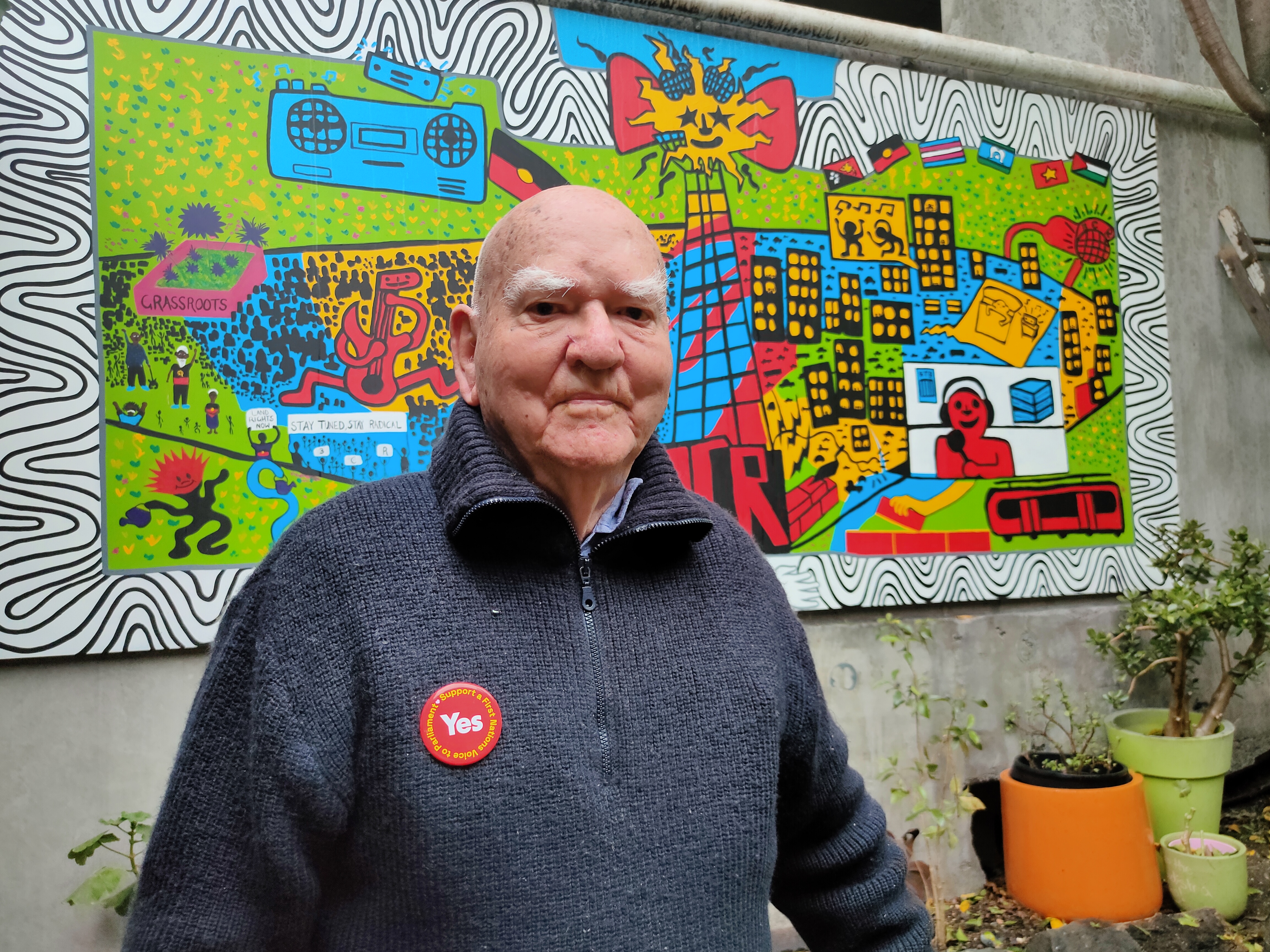 Percy stands in front of 3CR's new colourful mural in the courtyard. He wears a blue knitted jumper and sports a red YES badge