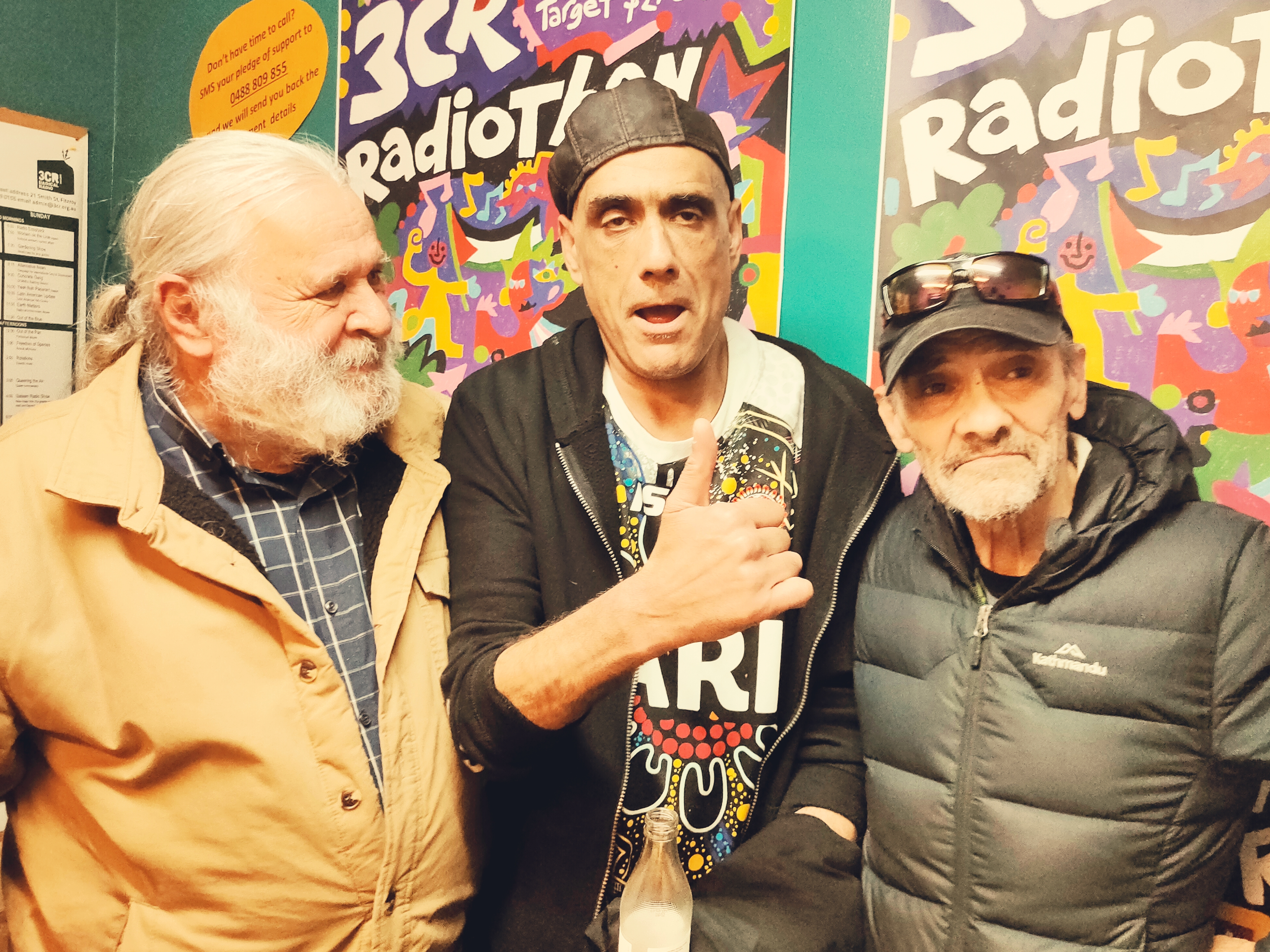 Robbie Thorpe stands next to fellow 3CR broadcaster, Gavin Moore, and host of Radical Australia, Joe Toscano. Robbie and and Gavin look straight ahead. Gavin gives a thumbs-up gesture. Joe looks sideways at Robbie and Gavin