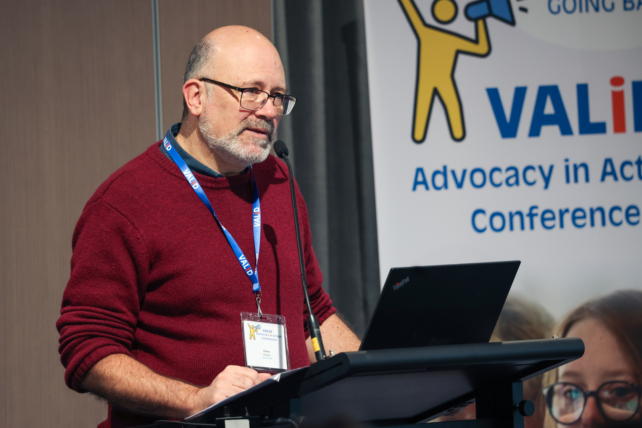 VALID CEO Fionn Skiotis, is standing at a podium and speaking into a microphone. He is wearing a red umper blue lanyard, and qlasses. Behind him is a white banner with vellow and blue graphics, text reads: 'VALID Advocacy in Action Conference'