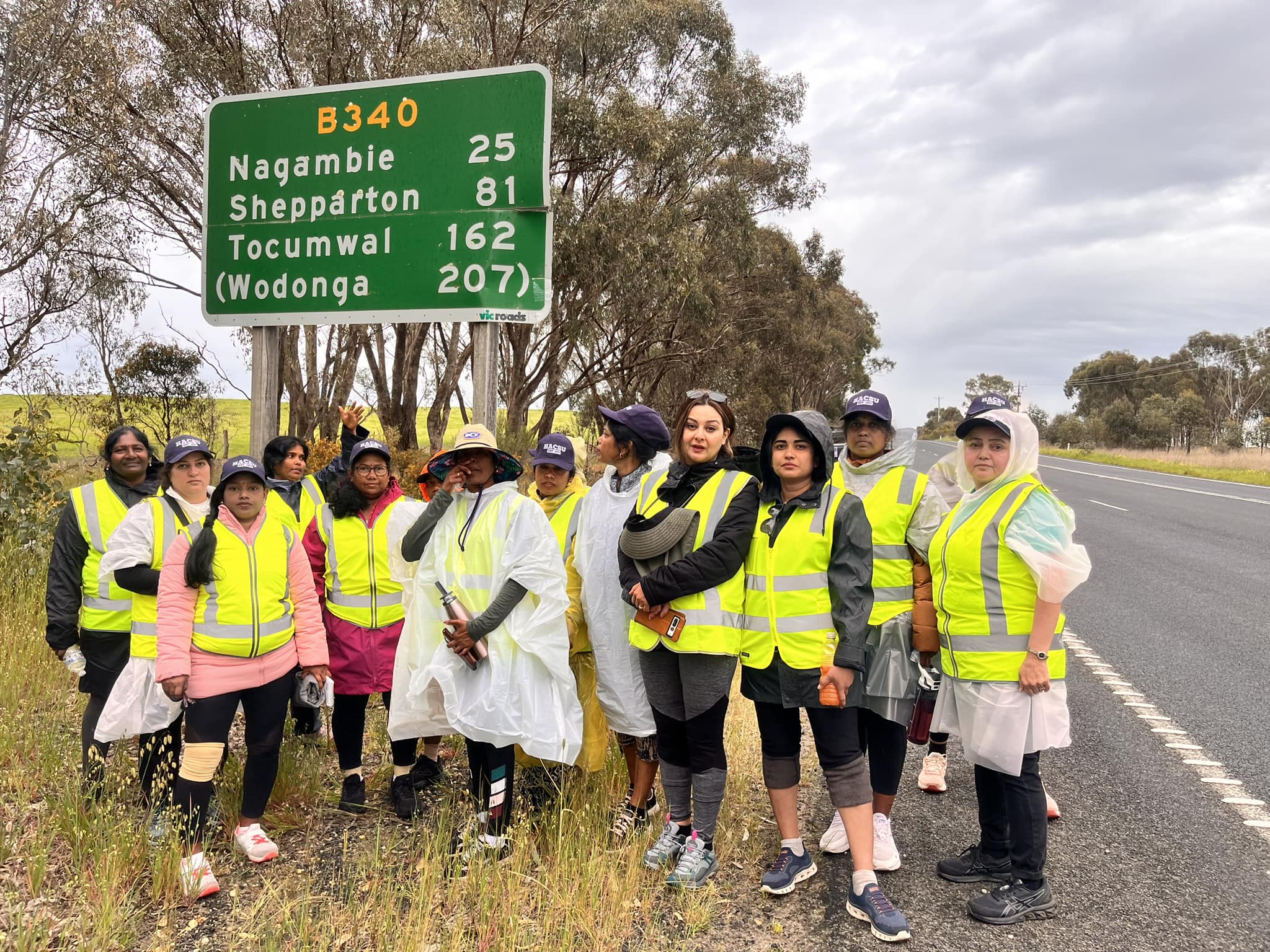 A photo of Refugee Women Action for Visa Equality wearing high vis vests, standing in front of a large road sign. They are standing on the side of a highway.