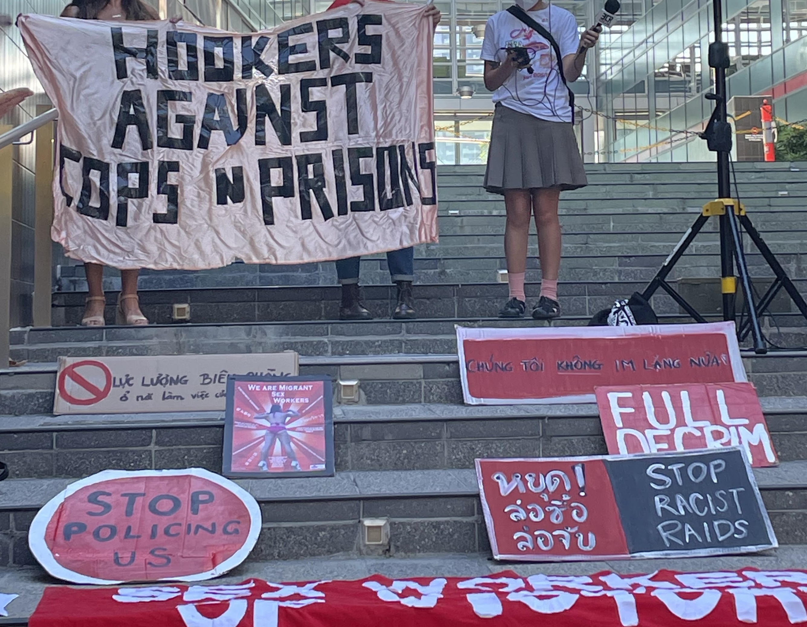 The foreground, steps in front of a corporate office building covered in handmade painted cardboard signs in various languages, two in English read: Stop policing us! and Full Decrim! The background shows two people holding a large homemade banner with black gaffer tape letters on light pink silky material that reads HOOKERS AGAINST COPS + PRISONS, with another person holding a microphone next to a speaker. The microphone has a little label on it that reads 3CR.