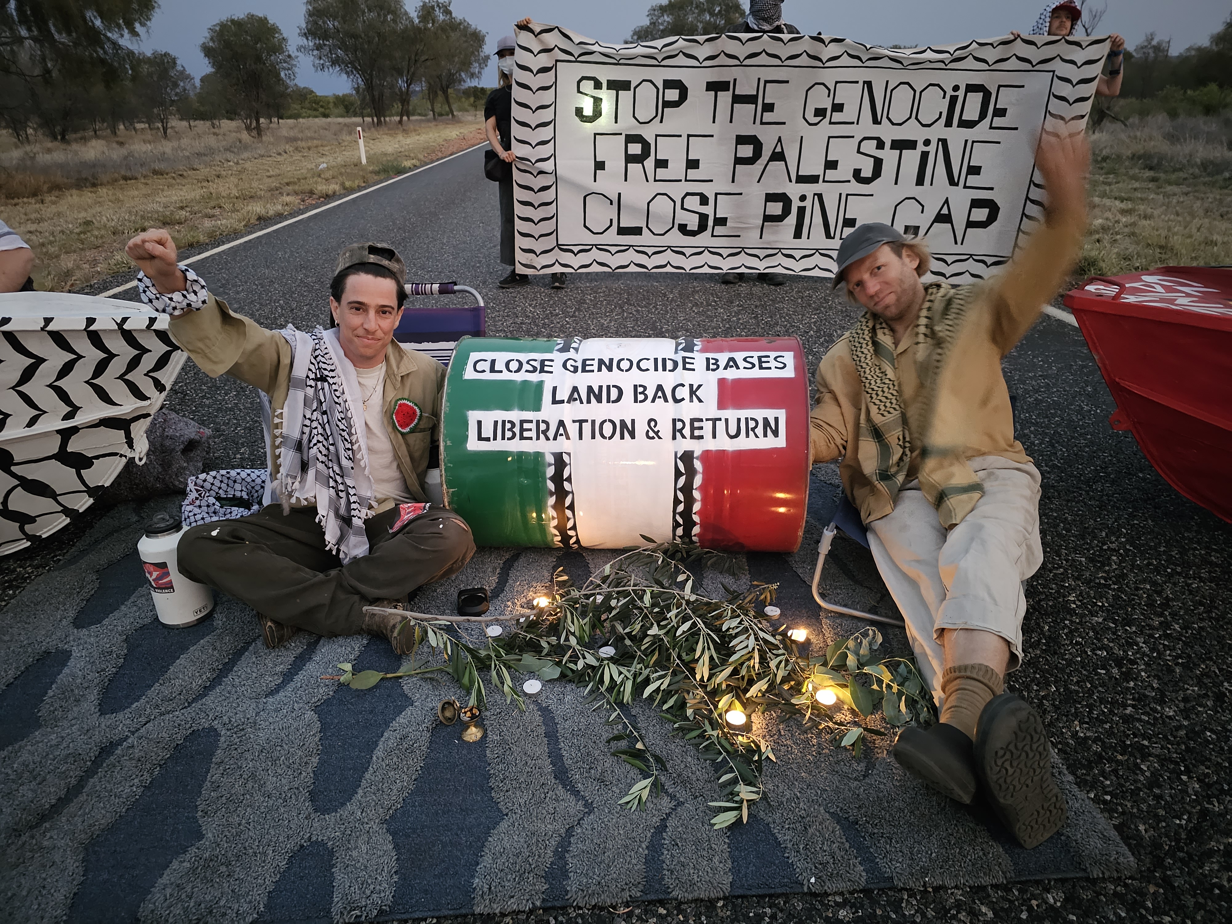 Jorgen Doyle and Yul Scarf sit on the access road to the Pine Gap military intelligence facility with a prop in colours of the Palestinian flag saying 'Close genocide bases, Land back, Liberation & Return'. Behind them, two people hold a banner saying, 'Stop the Genocide, Free Palestine, Close Pine Gap'.