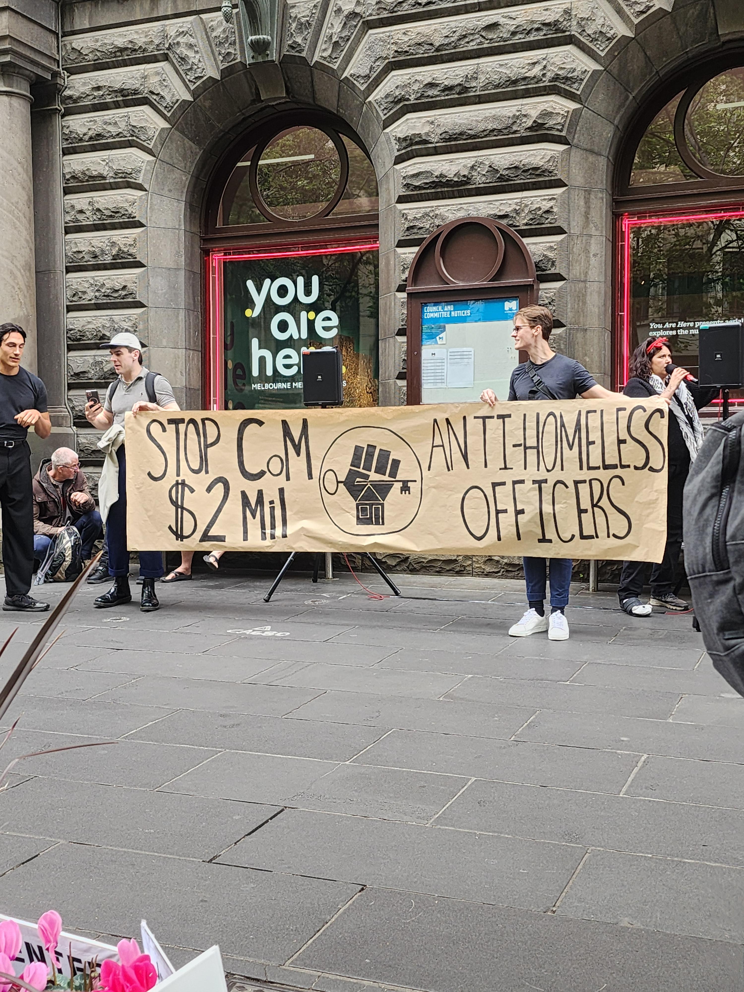 Two protestors hold a banner that reads 'Stop CoM $2m anti-homeless officers'. The logo of the Homeless Persons Union of Victoria is painted in the middle of the banner.