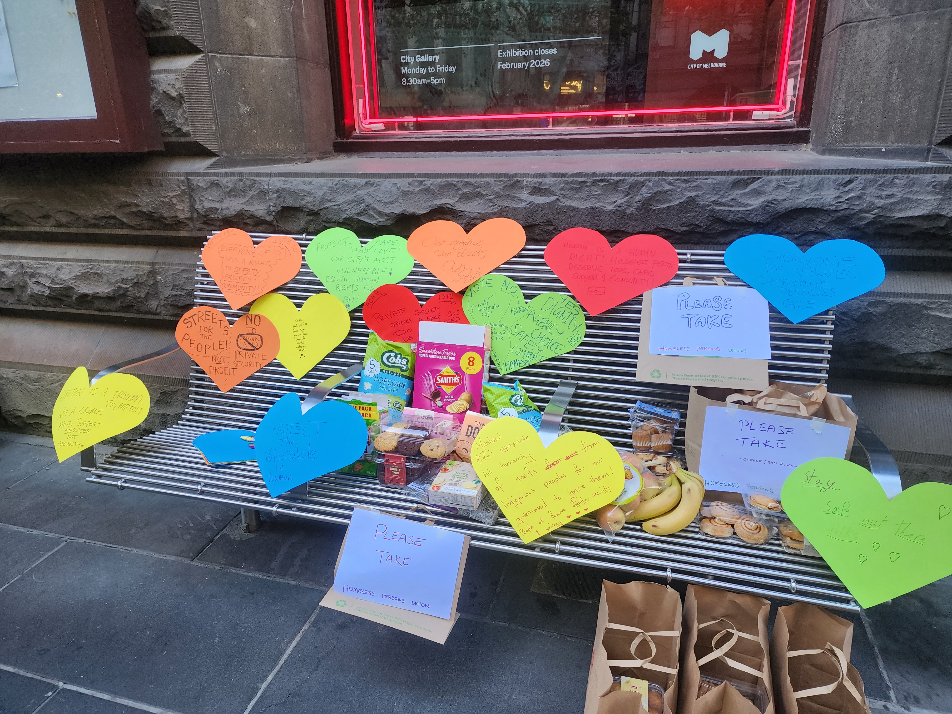 A city bench with large colourful paper hearts stuck all over it sharing messages of solidarity with Melbourne's homeless community. There is also an assortment of food on the bench.