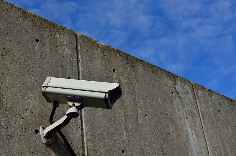 Surveillance camera on concrete wall facing downwards, with blue sky in the background. 