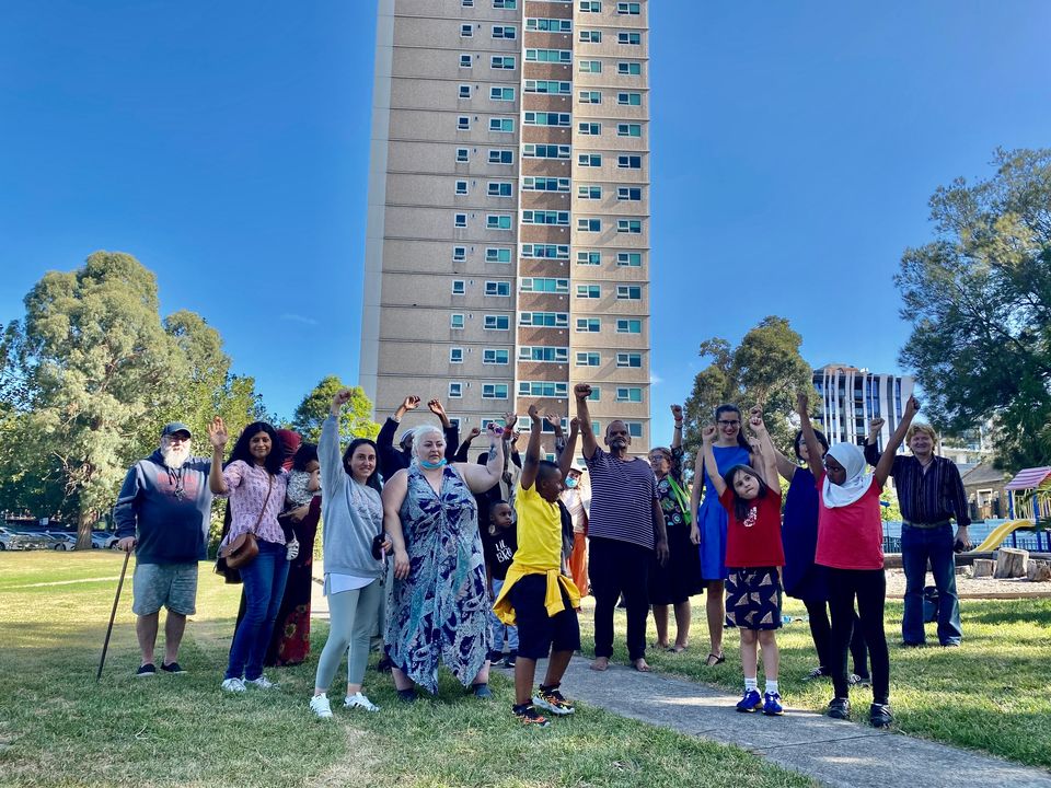 Collingwood public housing residents of a range of genders, ages, ethnicities and abilities stand together in the green space in front of one of the housing towers. Some of them raise their fists in the air. It is a bright and cloudless day.