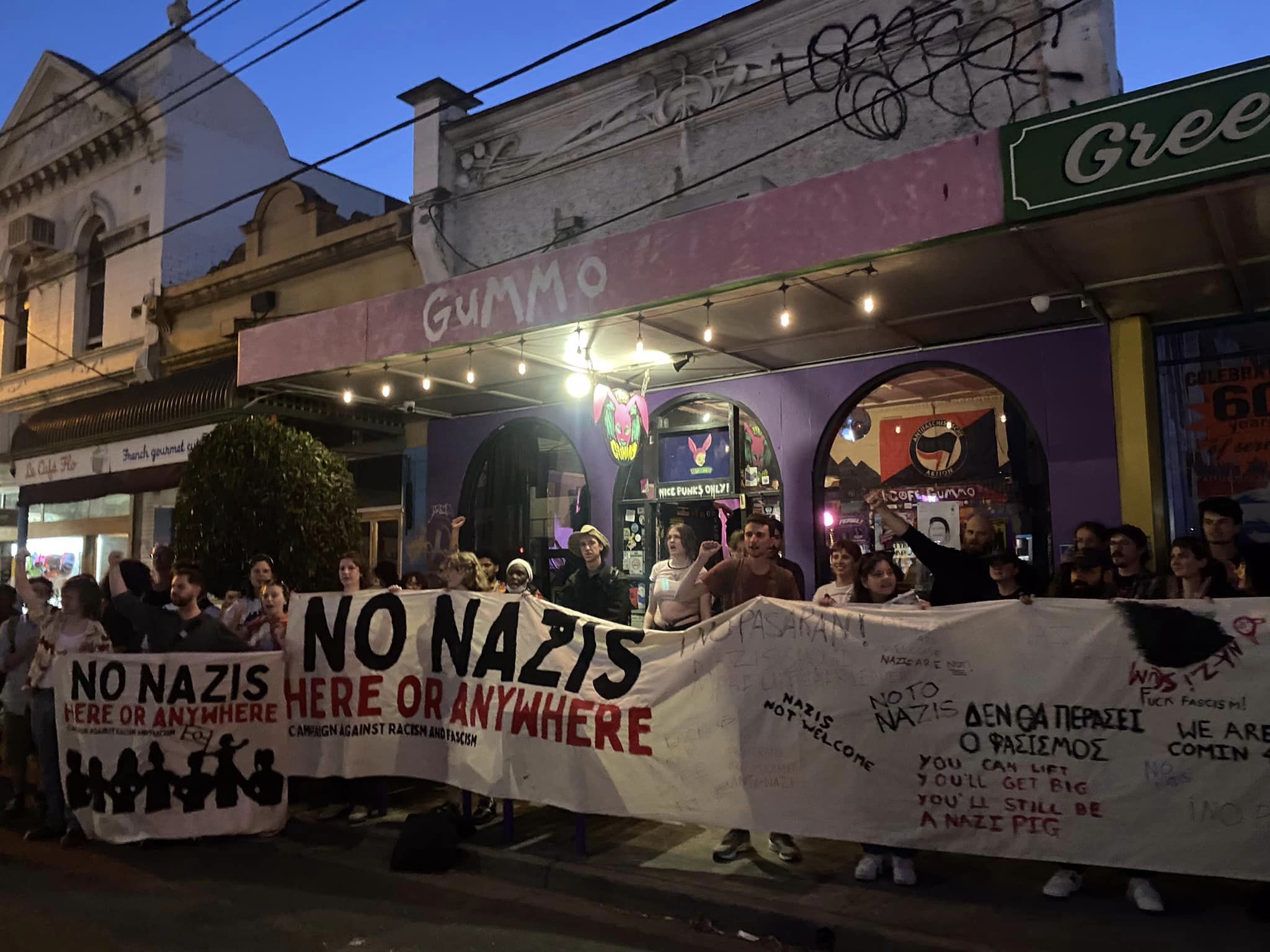 A photograph of a crowd of people standing on the pavement outside Cafe Gummo at dusk holding large banners that read 'No Nazis Here or Anywhere' and have anti-fascist messages in multiple languages painted across them.