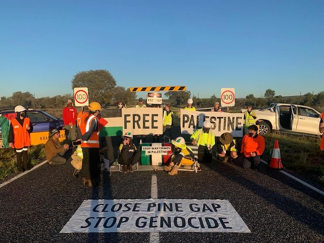 A photo of protesters blockading the Pine Gap access road in the early morning light. Two people are on the ground locked onto a barrel with a sign saying stop war crimes in Palestine, some are holding banners that say Free Palestine, and a large banner on the ground in front of the blockade reads Close Pine Gap Stop Genocide.
