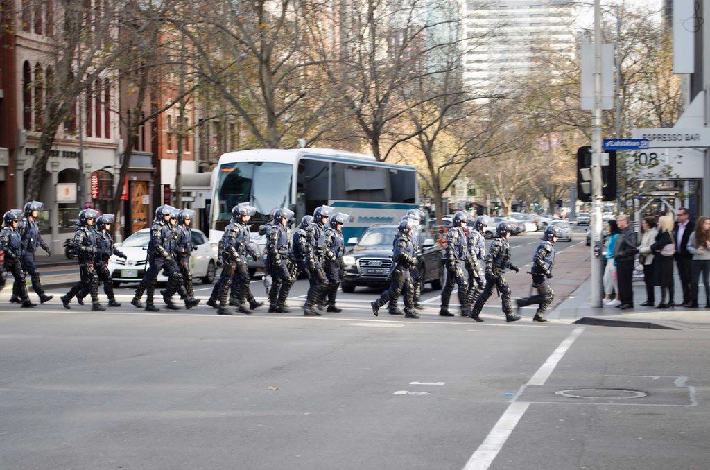 A photograph of members of Victoria Police Public Order Response Team crossing an intersection in single file, wearing full riot gear.