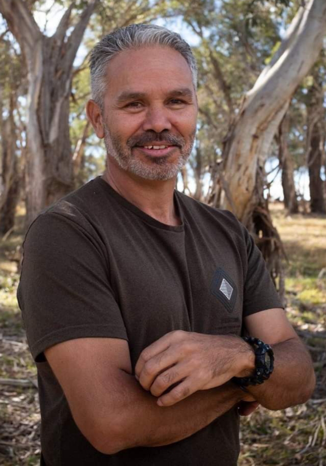 A man standing with his arms crossed and smiling with eucalypts in the background 