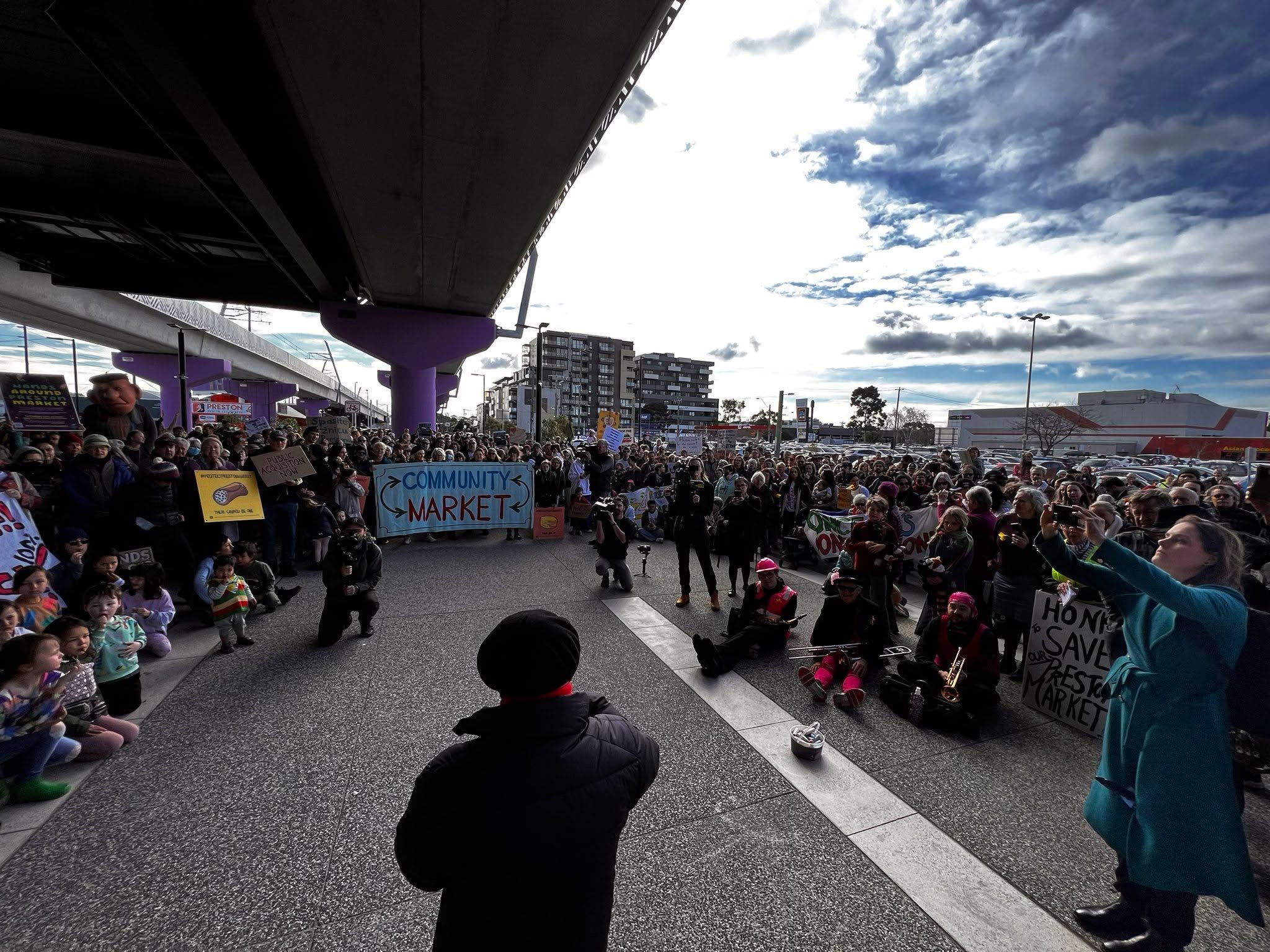 In the centre of the image we can see the back of Gaetano Greco as he addresses a gathering of people at the 2023 Hands Around Preston community protest to save the Preston Market from demolition and development. The gathering of people hold signs saying, 'Community Market,' and 'Honk to save our Preston Market'. 