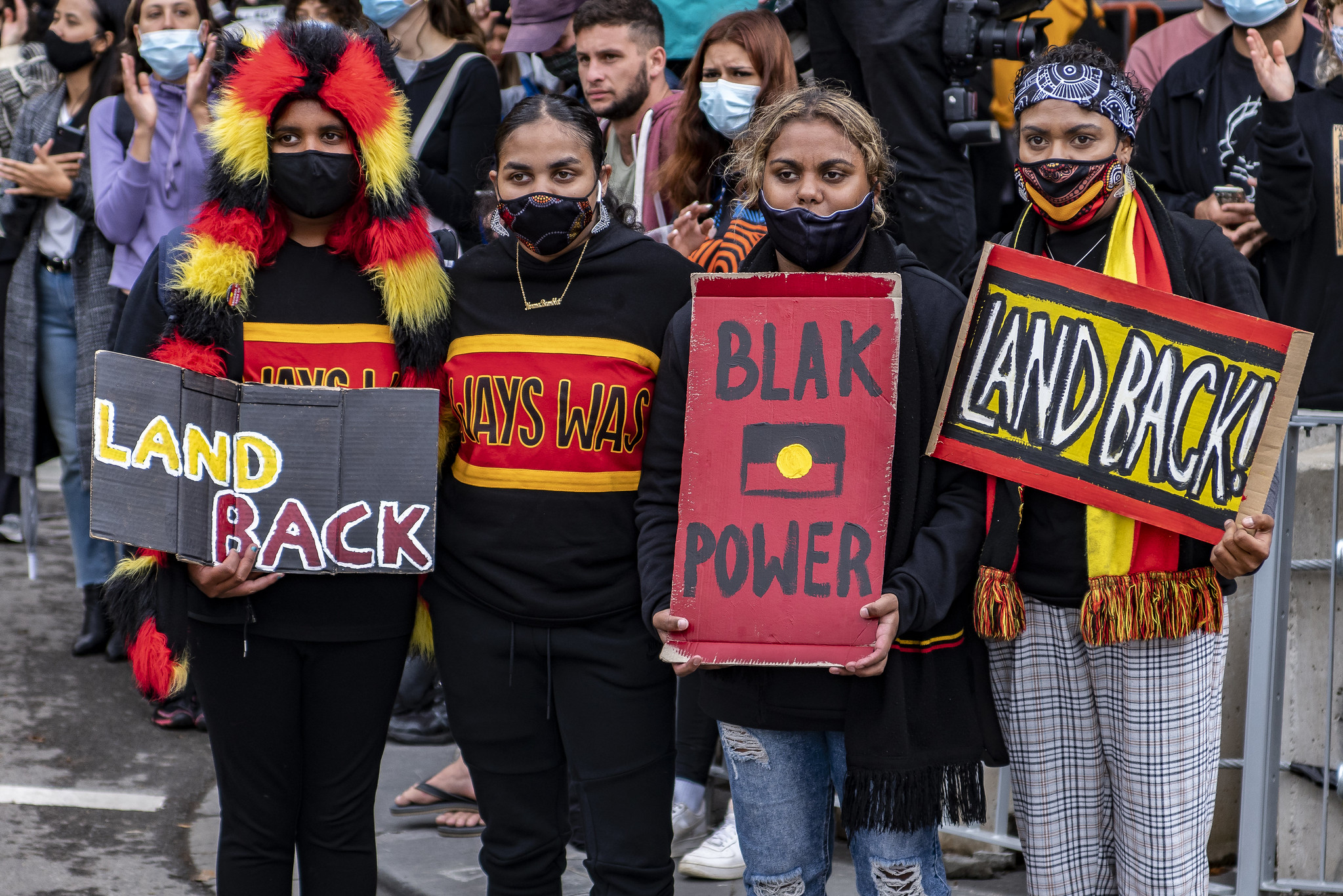 A photograph of four young Aboriginal women at the Invasion Day 2021 rally in Melbourne holding signs that read Land Back and Blak Power.