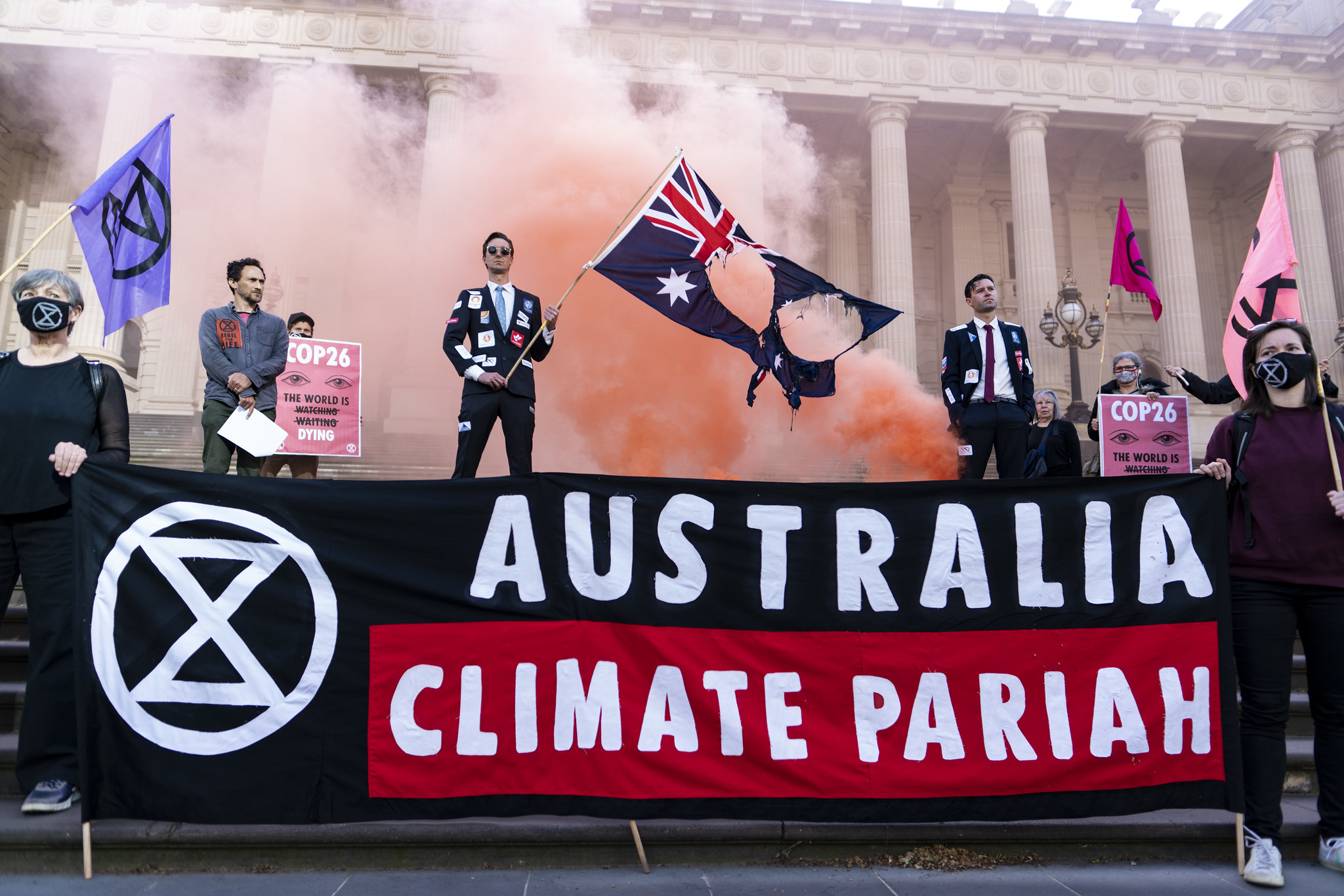 A photograph of a group of Extinction Rebellion activists on Parliament House steps in Melbourne. Two stand at the bottom holding a large banner bearing the XR symbol and which reads "Australia, Climate Pariah". Two men in suits covered with stickers from extractive industries companies stand behind the banner, and one is holding an Australian flag with holes burned in it. Others stand on the steps holding signs and XR flags. Orange smoke fills the background.
