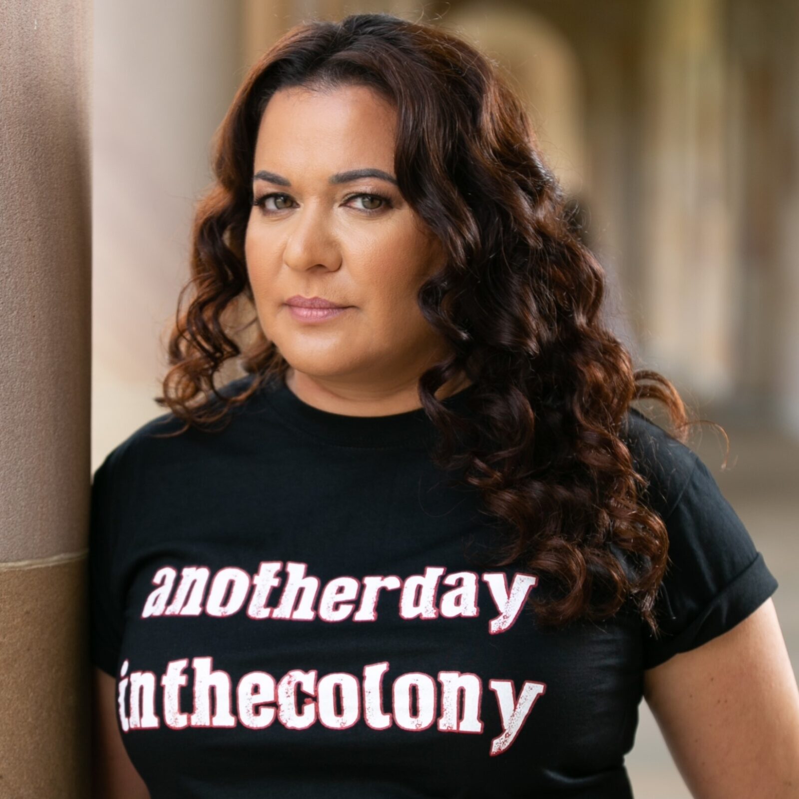 A photograph of Chelsea Watego standing beside a sandstone pillar in an open walkway. She wears a black t-shirt printed with the words 'another day in the colony' printed on it in white lettering with red borders, and she is looking at the camera with a serious expression. Chelsea's hair is dark brown and curly, and is worn down, falling across her left shoulder.