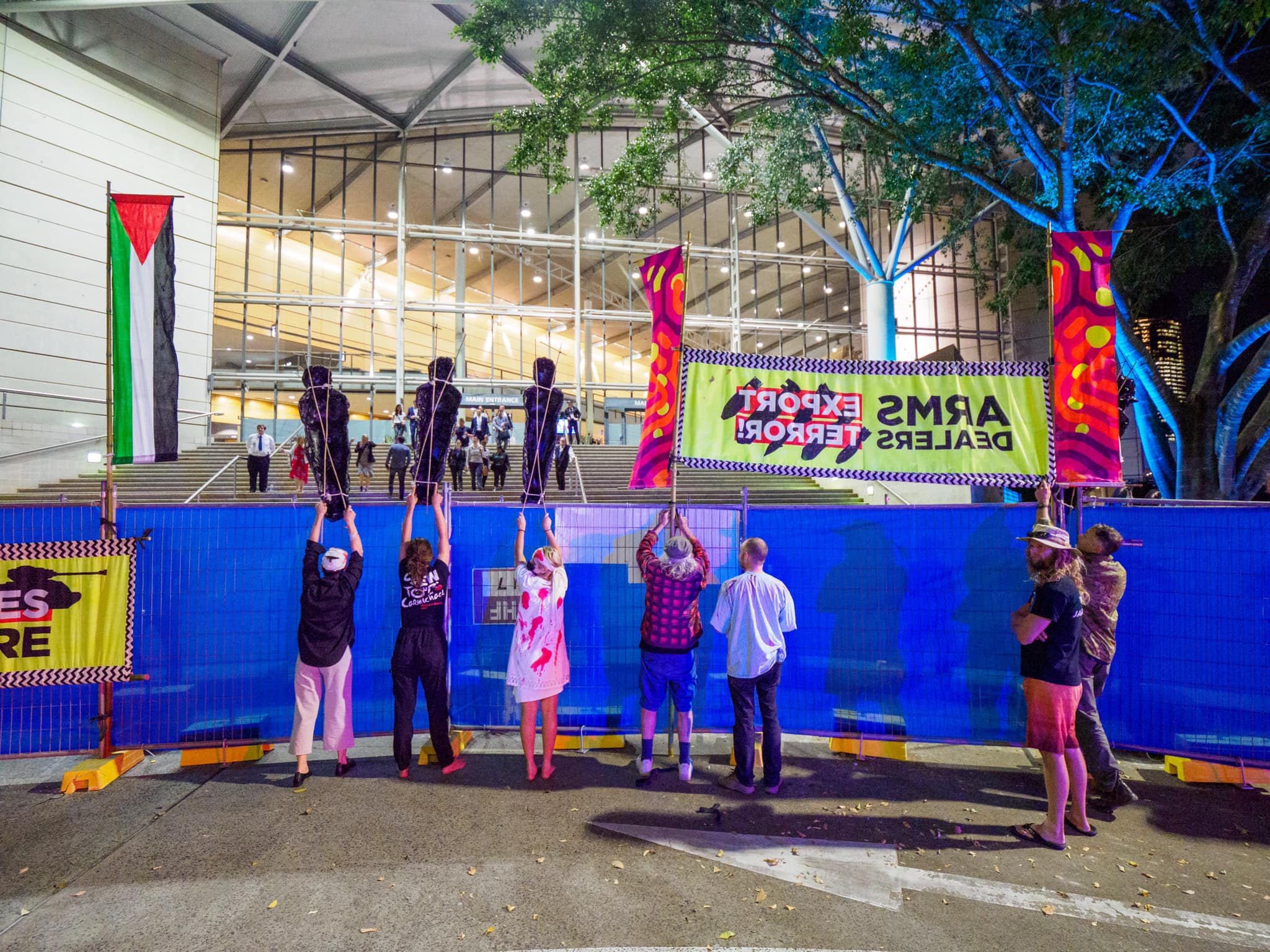 A photograph of protestors peacefully demonstrating outside the Brisbane Convention Centre where Land Forces is being held. Protestors are prevented from getting closer by a tall blue barricade, but they hold up black fibreglass casts of the figures of children and a colourful banner that reads 'Arms Dealers Export Terror!' A Palestinian flag hangs vertically to the left of the protestors.