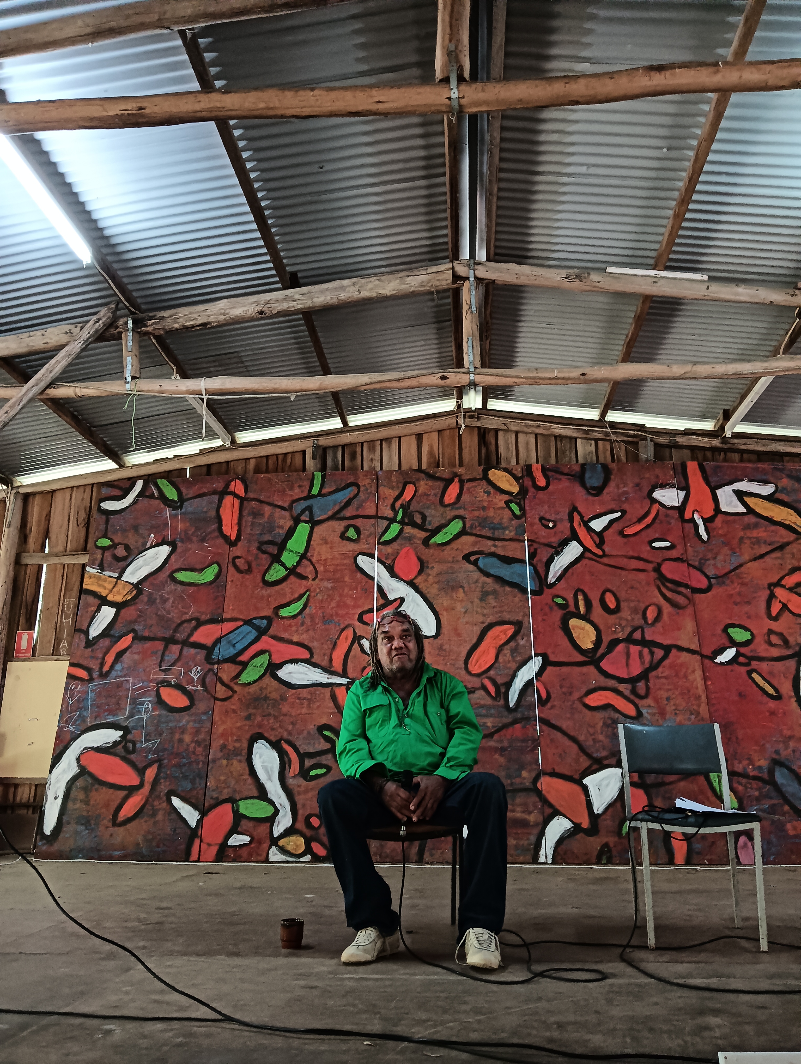 Wayne Coco Wharton sits on a stage in a large open shed with a multicoloured mural in the background. He is talking and wearing a green long sleeved shirt and black pants.