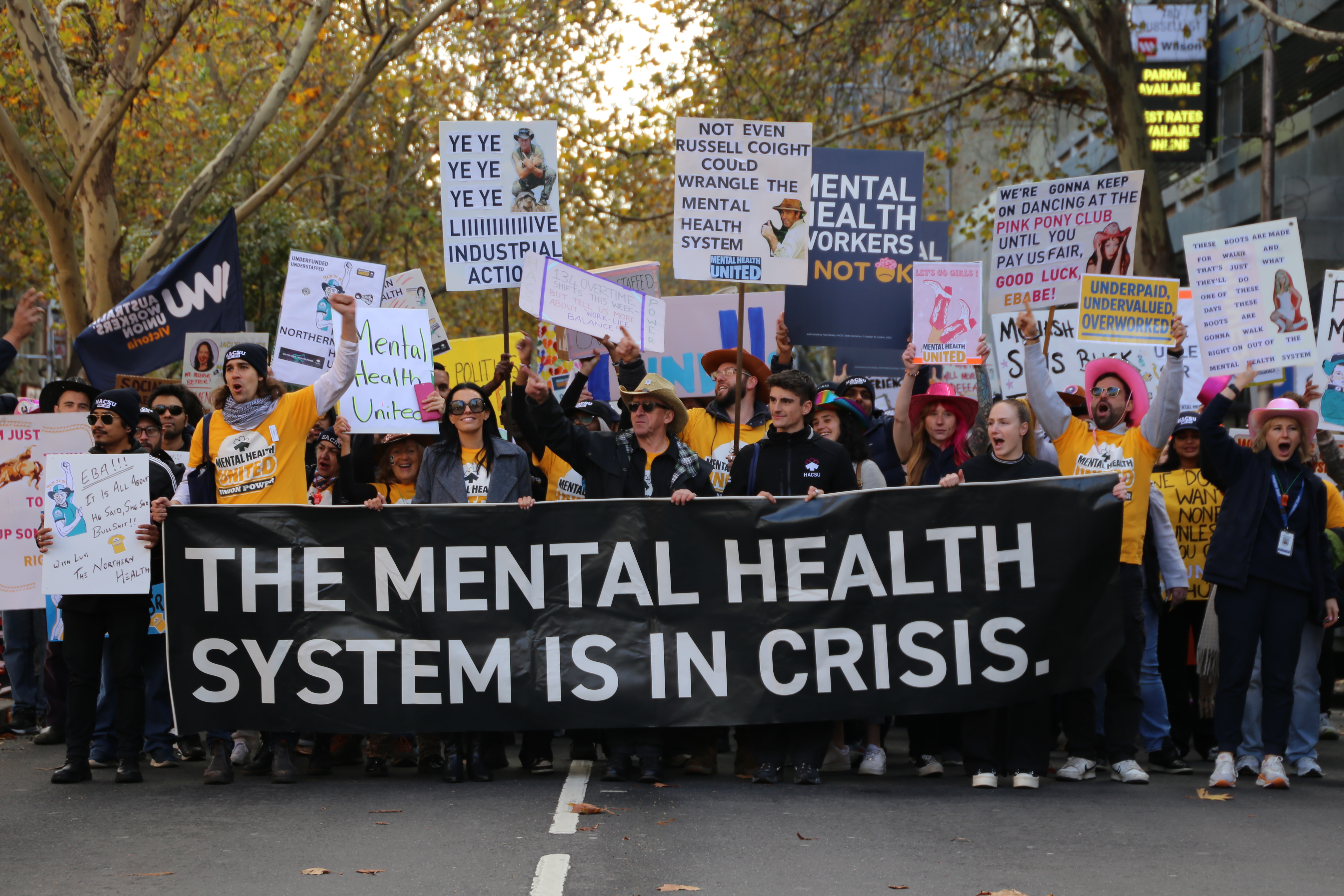 HACSU Rally Tuesday 17th of June. Black banner with white bold font reads 'The Mental Health System is in Crisis'. Rally members in yellow and black holding up signs and chanting. 