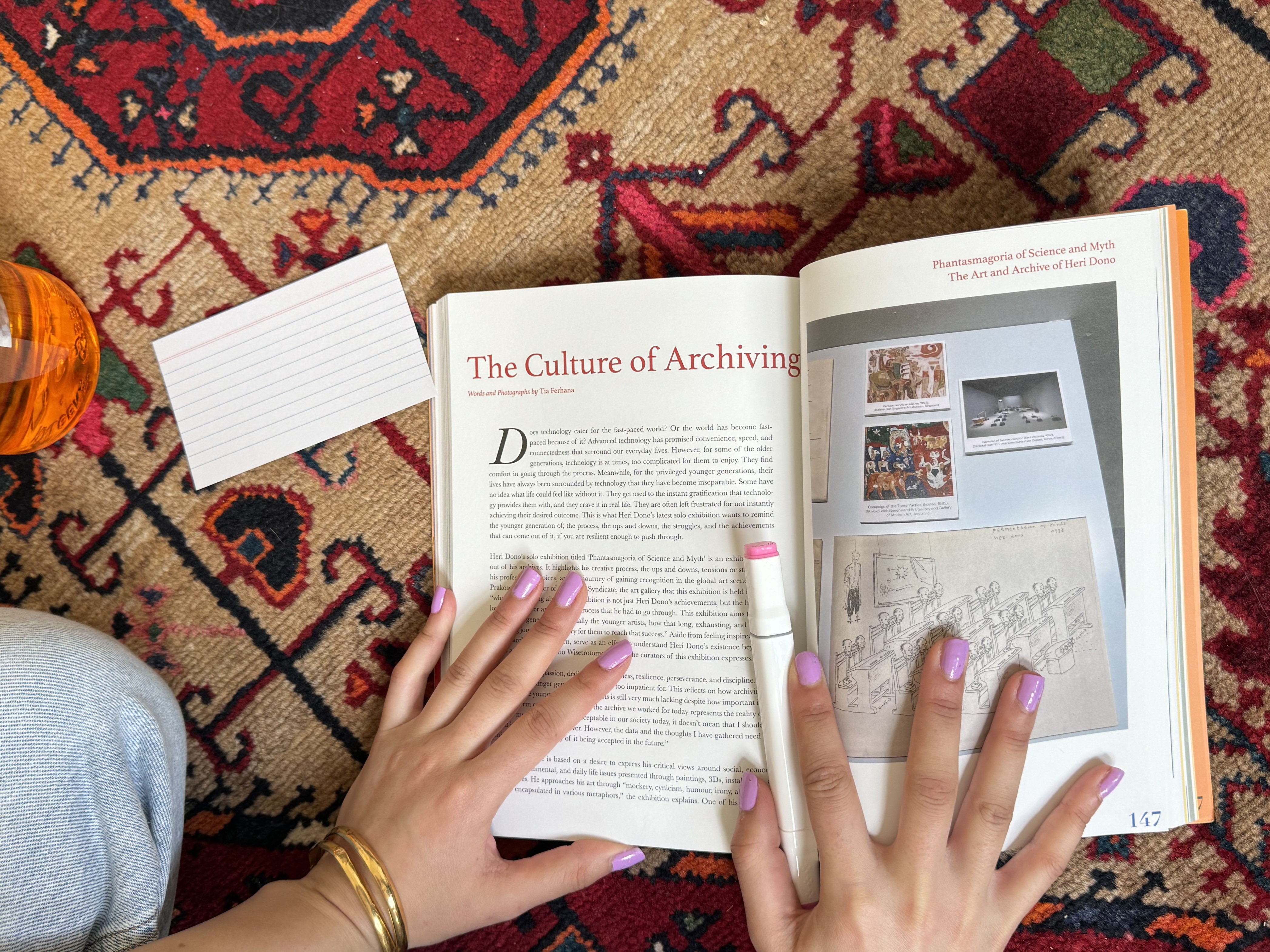 The Great Book Return. Open face book reading The Culture of Archiving with picture on right hand side, being held open by hands with light pink nails holding pink highlighter wearing gold bracelet on left hand. Blank lined cue cards lay next to the book, with background red, white rug with intricate lines and shapes. 