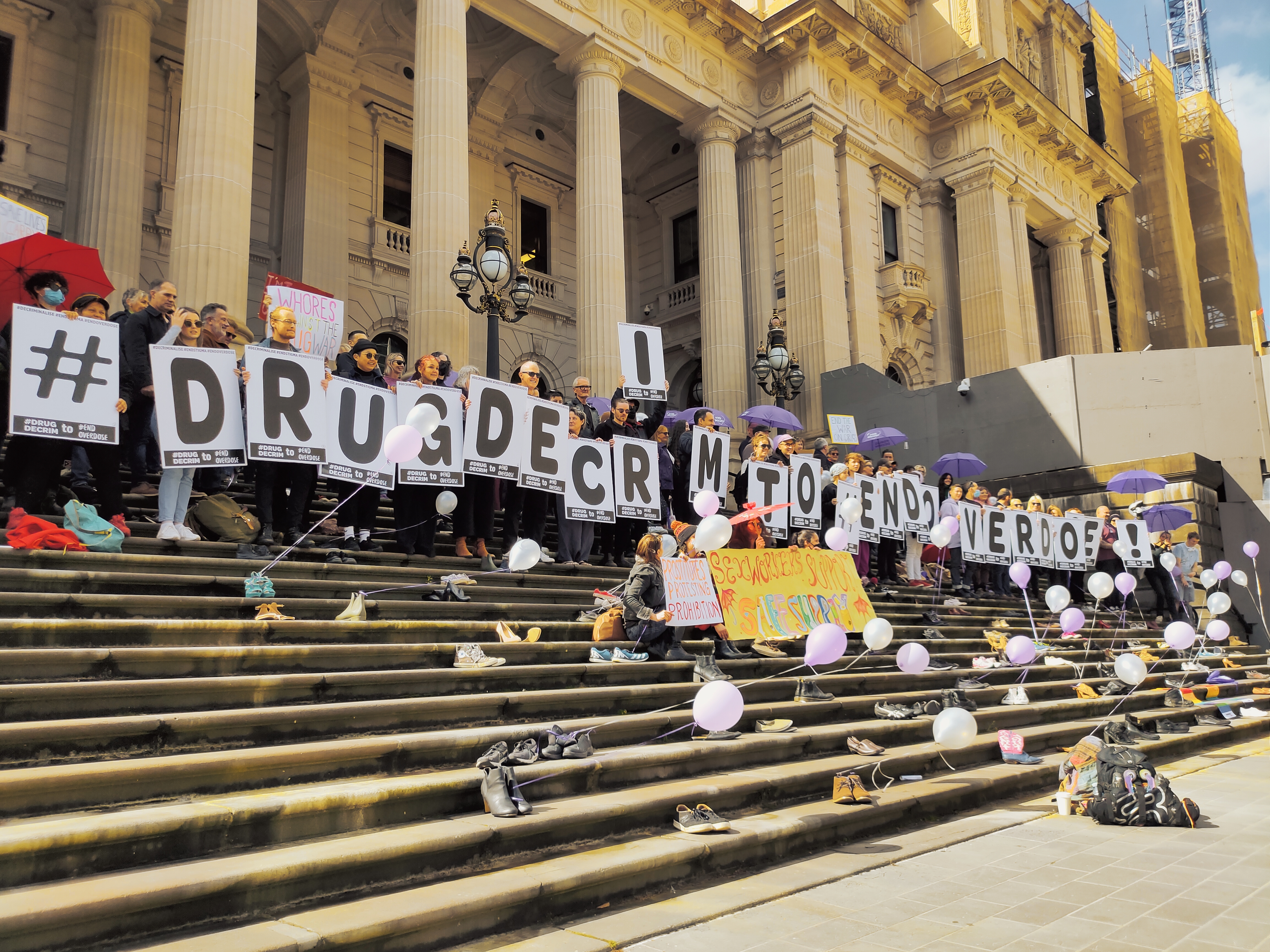 A crowd of people gather on the steps of Parliament House holding signs with individual letters that spell out "# DRUG DECRIM TO END OVERDOSE".