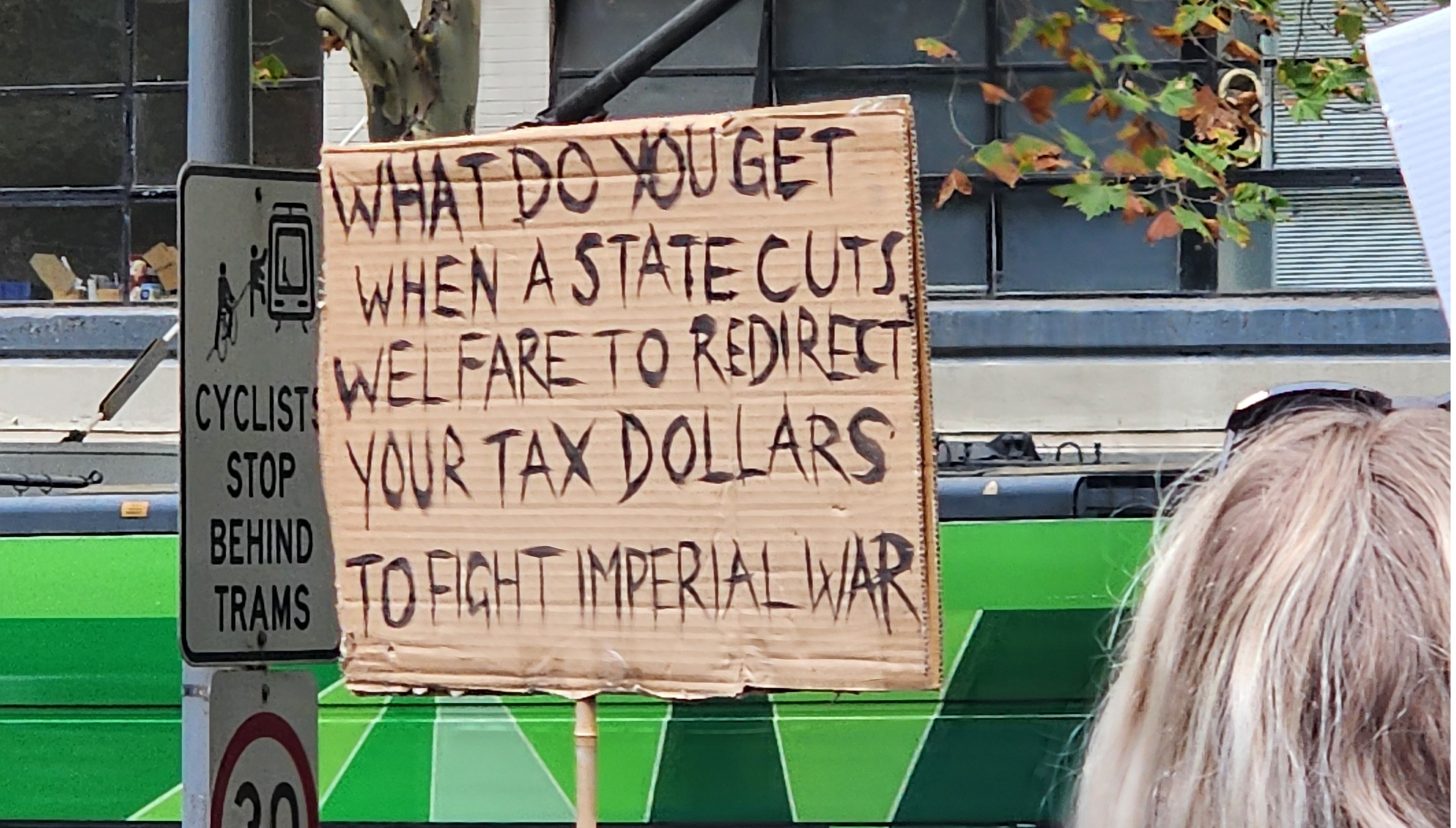 Photograph of a handwritten carboard protest sign at the NDIS rally in Naarm 2026 reading "WHAT DO YOU GET WHEN A STATE CUTS WELFARE TO REDIRECT YOUR TAX DOLLARS TO FIGHT IMPERIAL WAR".