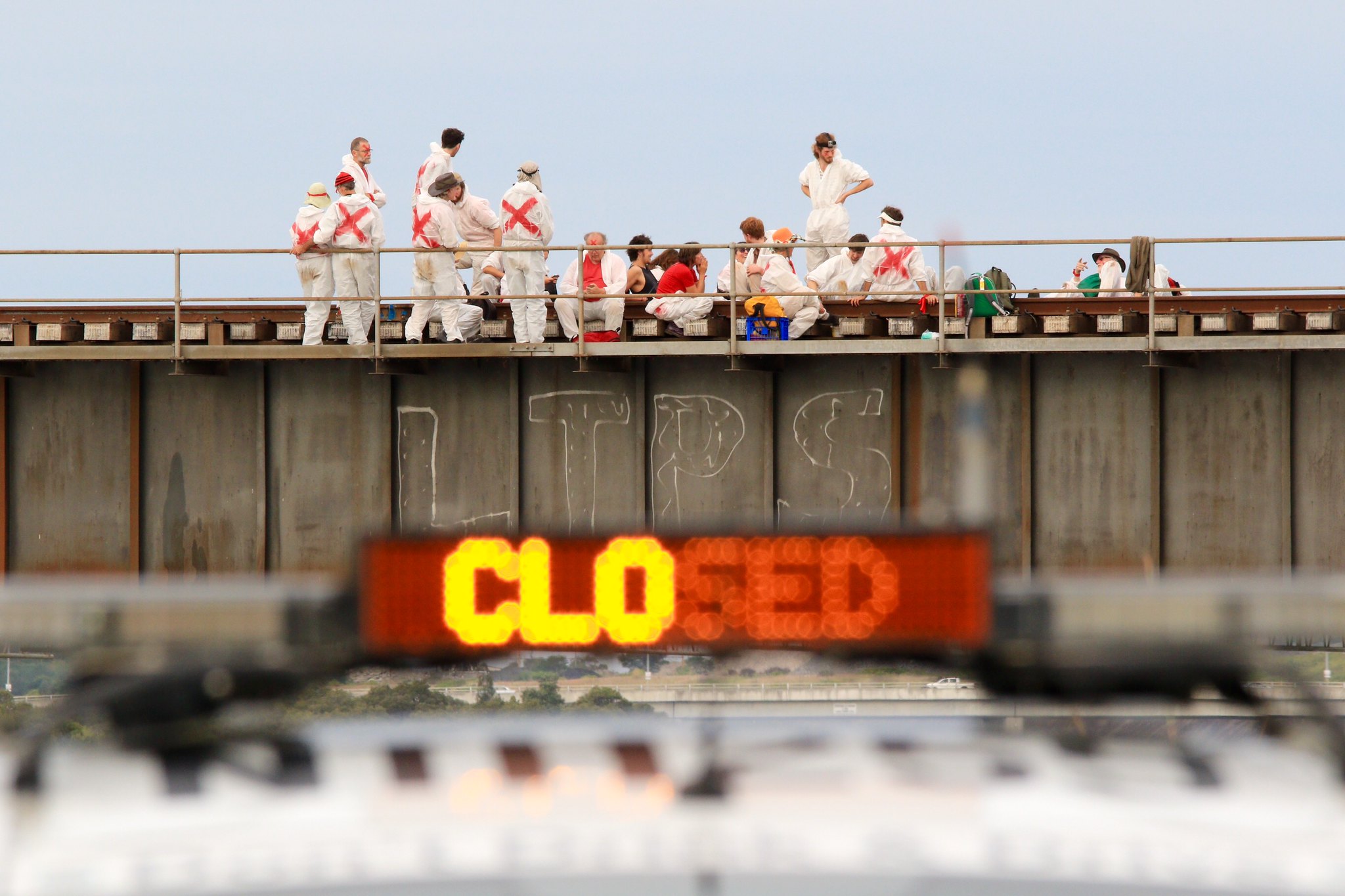A photograph of protesters dressed in white jumpsuits with red 'x' on the back blocking a railway bridge in Newcastle. A sign on the side of the bridge reads 'CLOSED'.