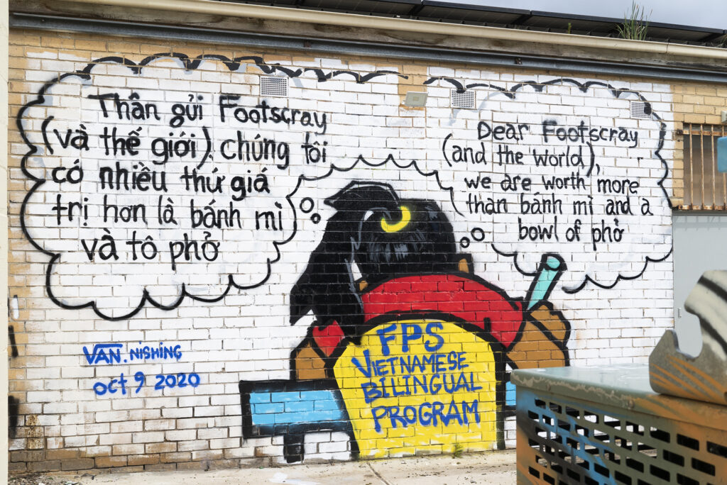 A piece of graffiti art on a wall depicting a student with long black hair in a ponytail, a red shirt and tan skin sitting at a desk and writing. The student is shown from behind, and the back of their chair reads 'FPS Vietnamese Bilingual Program'. They have two thought bubbles, one in Vietnamese and one with the English translation 'Dear Footscray (and the world), we are worth more than bánh mì and a bowl of phở'.