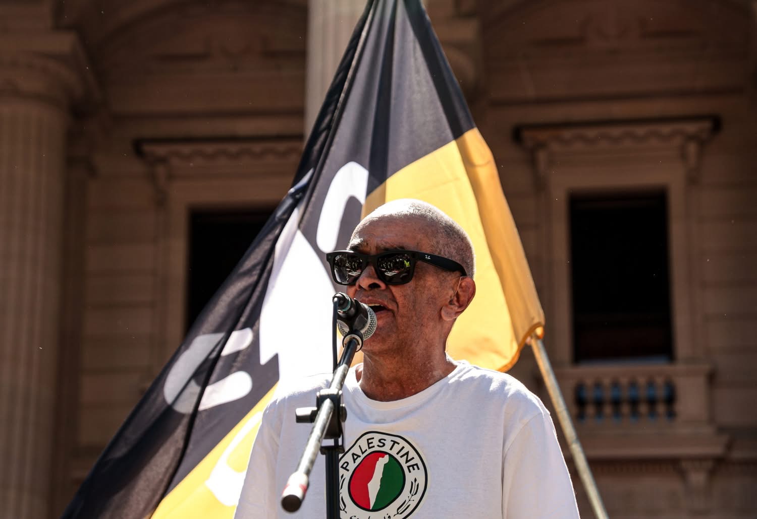 Professor Gary Foley speaking at the Naarm Invasion Day Rally 2026. He is wearing a shirt with a map of Palestine and the Camp Sovereignty flag flying behind him.