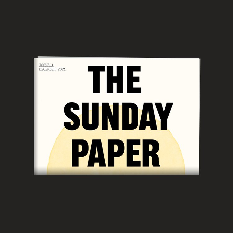 A digital image of The Sunday Paper, a folded newspaper on a black background. The cover has 'The Sunday Paper' written across it in large black capital letters, with half a yellow painted sun rising from the fold behind the text. In the top left corner is printed 'Issue 1 December 2021'.