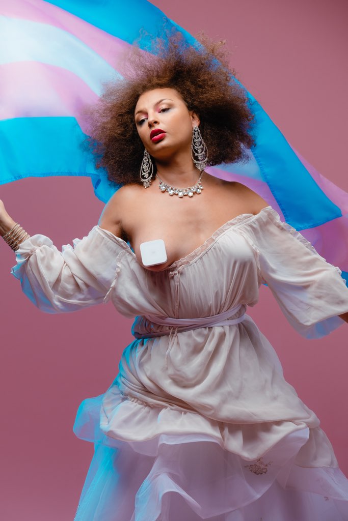 A photograph of Miss Cairo wearing a ruffled, flowy pale pink dress pulled down in front to reveal one censored breast. She holds the trans flag up with one hand, and it waves behind her head. She is wearing elaborate crystal earrings, a necklace and a bracelet.