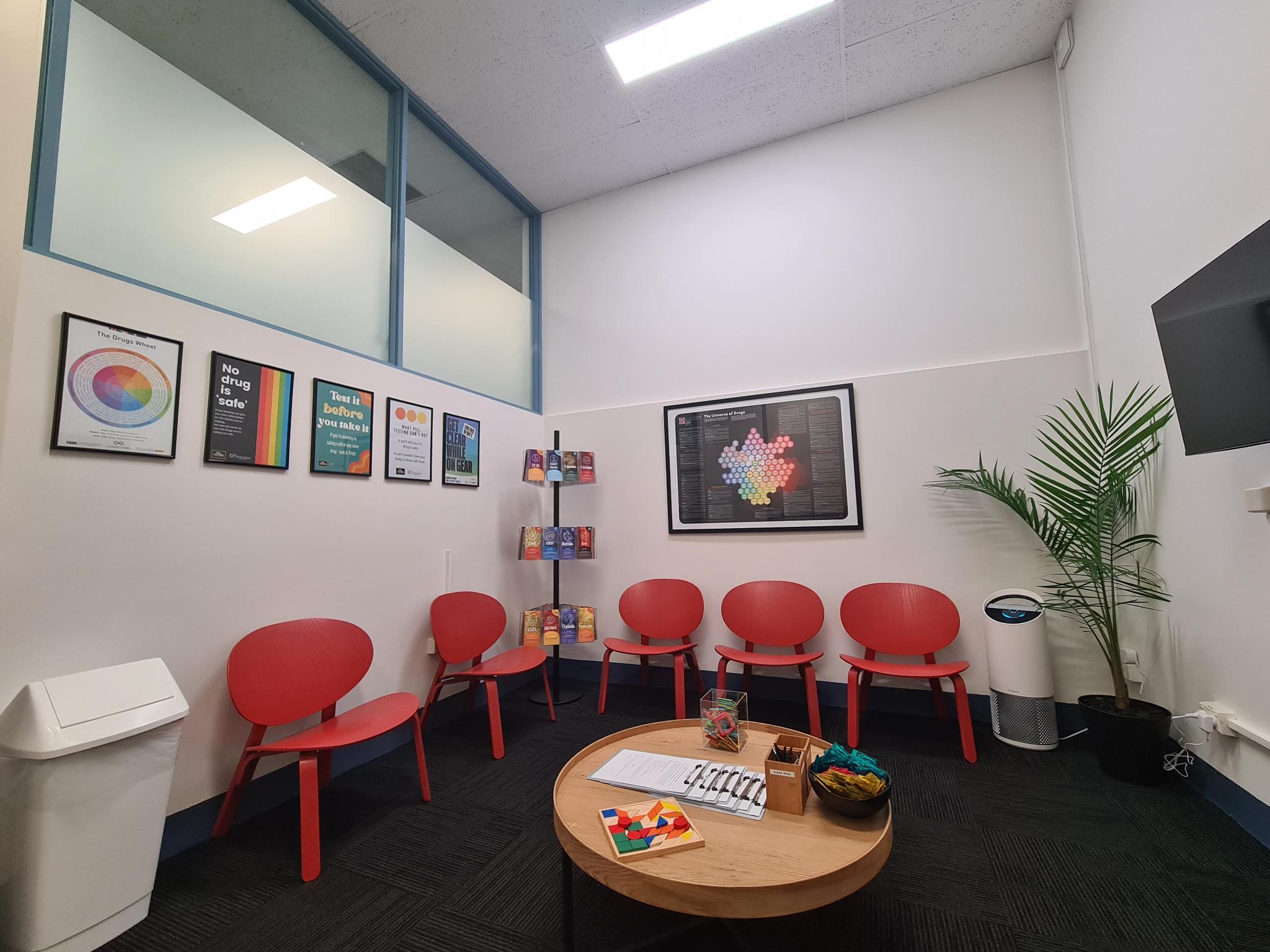 A photograph of the CanTEST Health and Drug Checking service waiting room. The room has red chairs lining the wall, a low table, and posters on the wall with health and harm minimisation information about drugs.