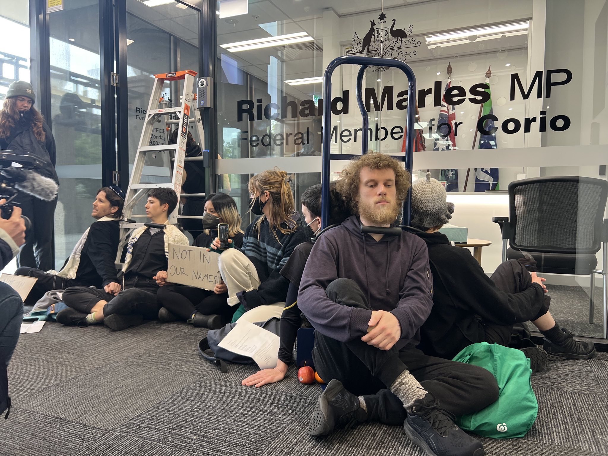 A group of protesters sit on the floor outside Richard Marles' office. Some of them are locked on by the neck to a trolley, while others are locked on by the neck to a ladder standing in the open doorway of the office.