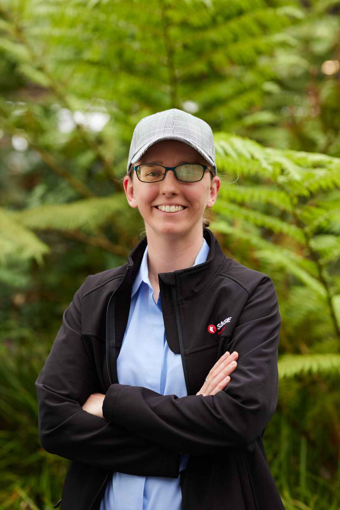 Cassie Hames is wearing a cap and black jacket saying Sage Automation with her arms folded with a content smile on her face standing in front of Green plants.