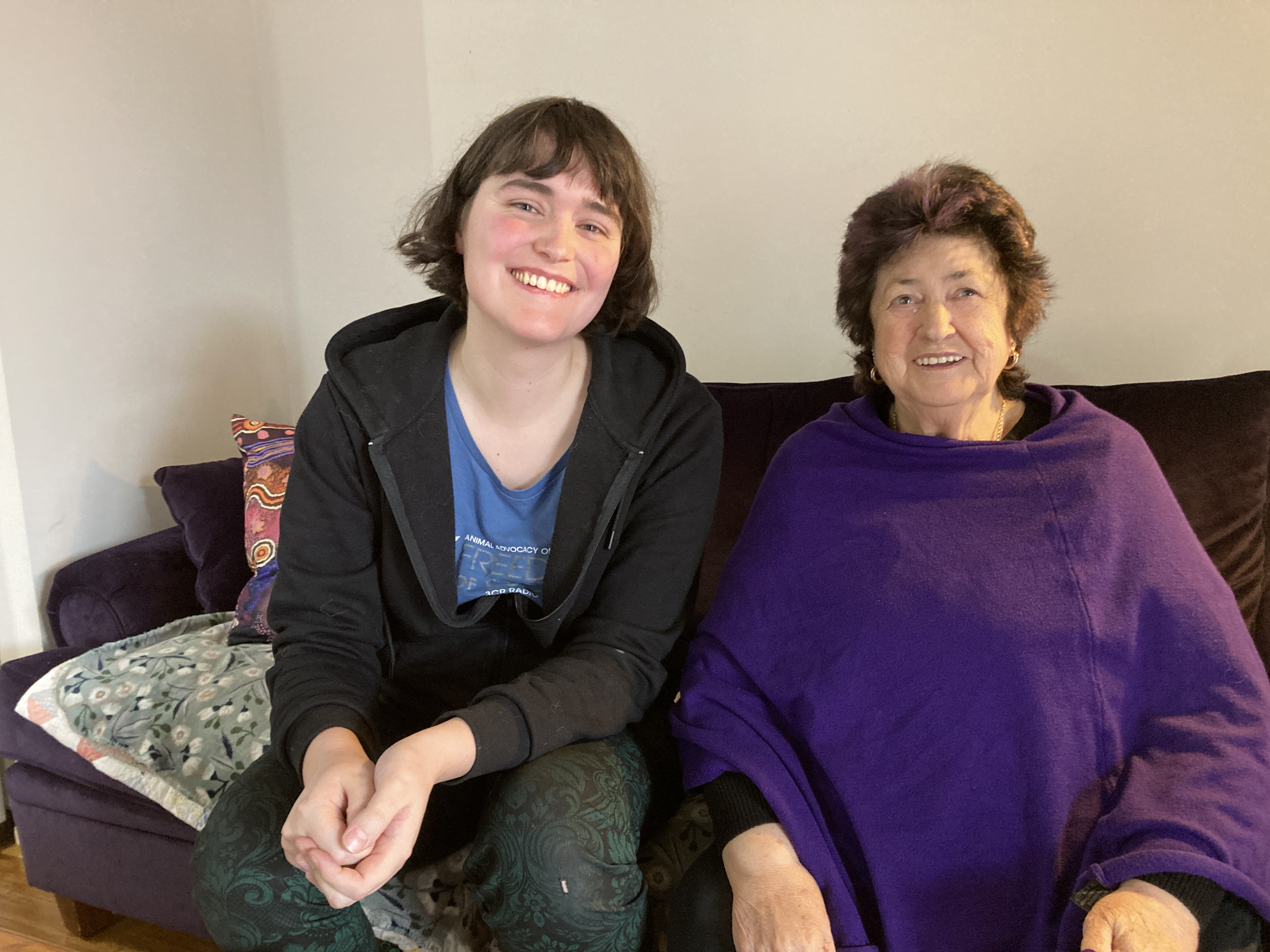 Amy and Aunty Lyn sitting next to each other on a couch smilling for a photo graph. Amy has an opened, black zipper hoodie with a 3CR T-Shirt. She has brown hair in a bob with bangs. Aunty Lyn is smiling away in a purple poncho with short dark hair with light purple hair dye. 