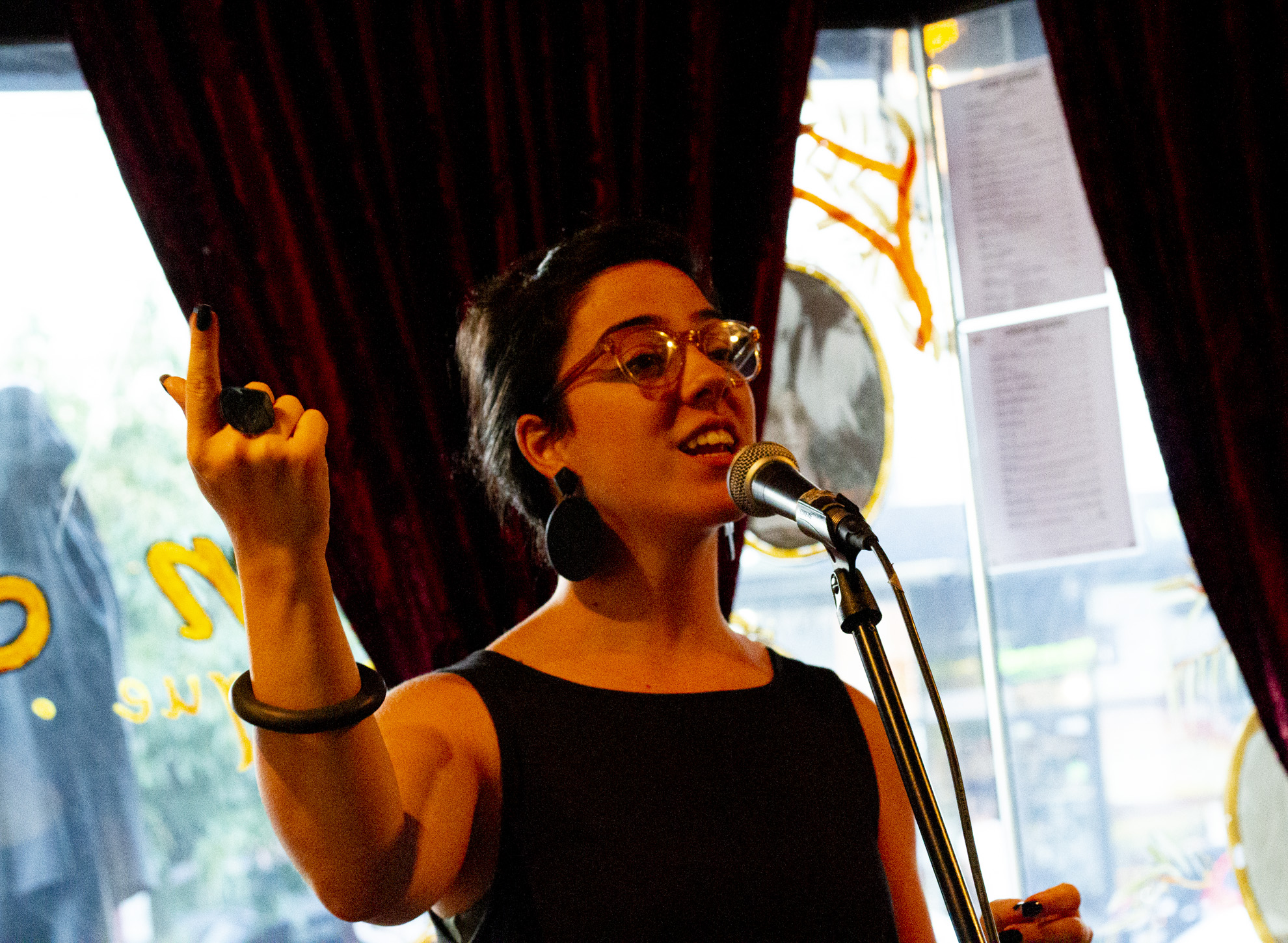 Ren Alessandra, young woman speaking live behind a microphone, one hand gesturing expressively