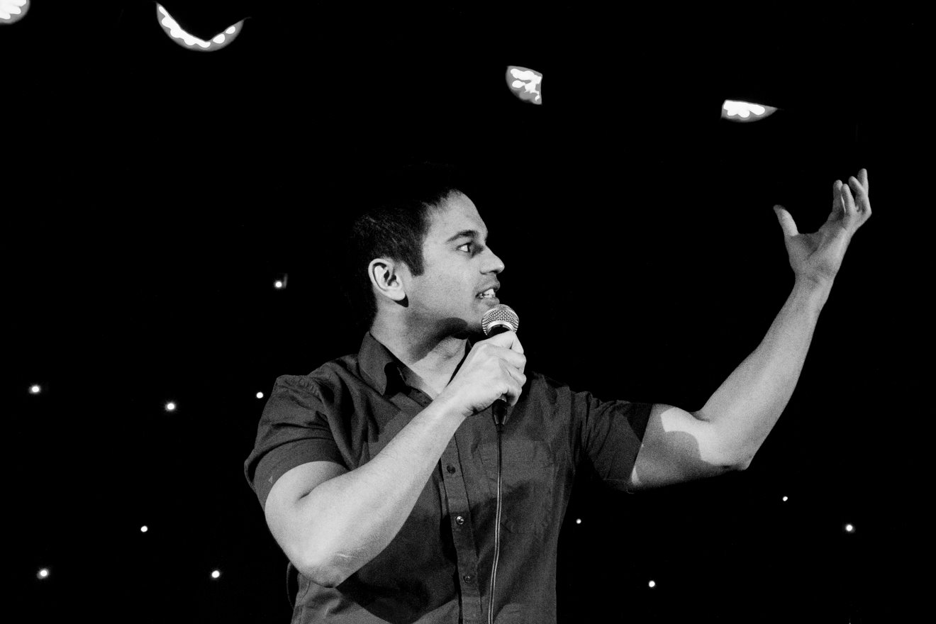 Medium close up of a male poet, Josh Cake, performing on stage. He is holding a microphone in one hand and gesturing expressively with the other 