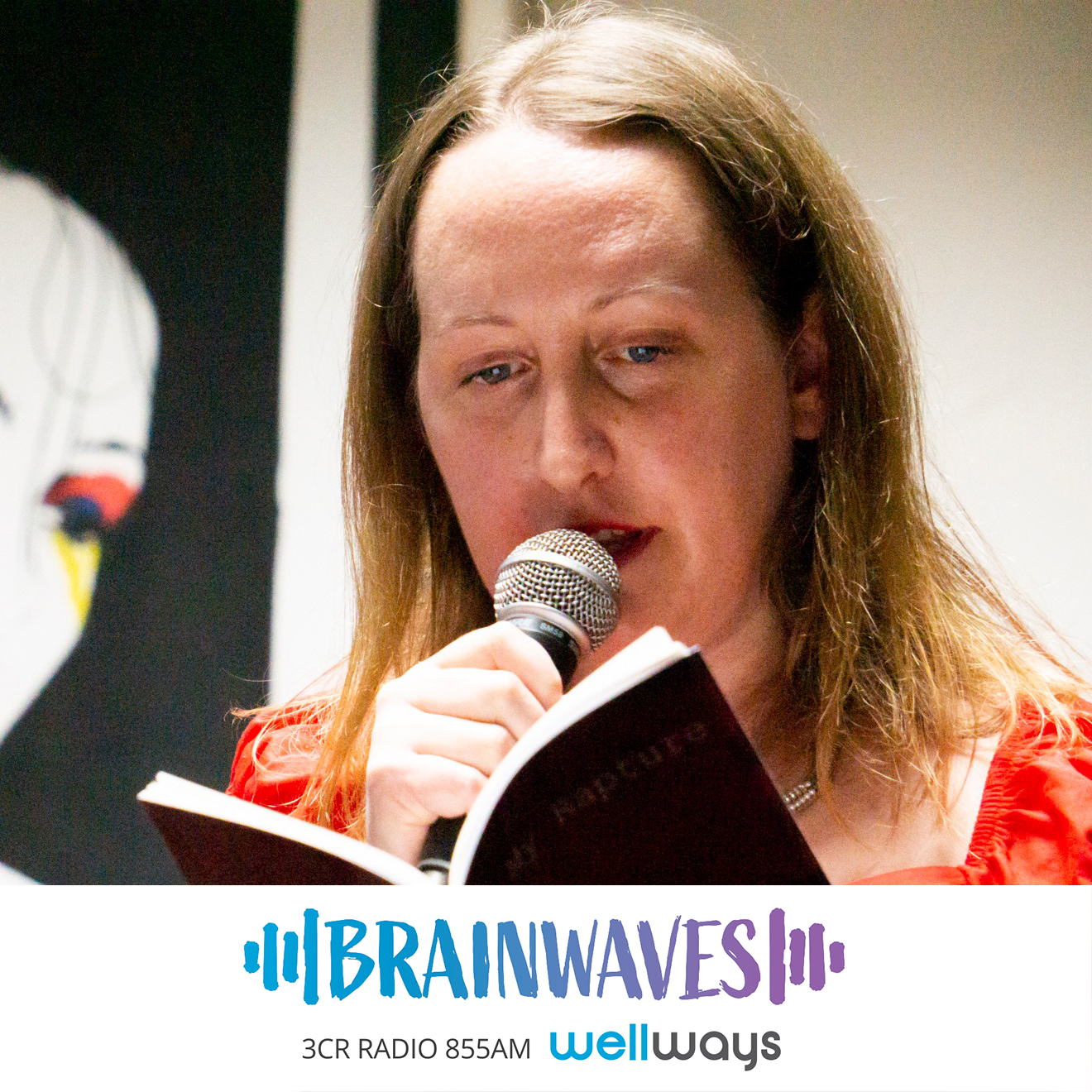 Gemma White is photographed reading her poetry at a live event. She is a caucasian woman with blonde hair wearing a red top, holding a microphone to her mouth as she reads from a book with a black cover. There is a large piece of wall art behind her.
