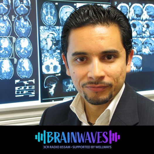 A man with dark hair stands in front of some x-rays of brains. Brainwaves Logo in blue and purple is below the image in a banner.
