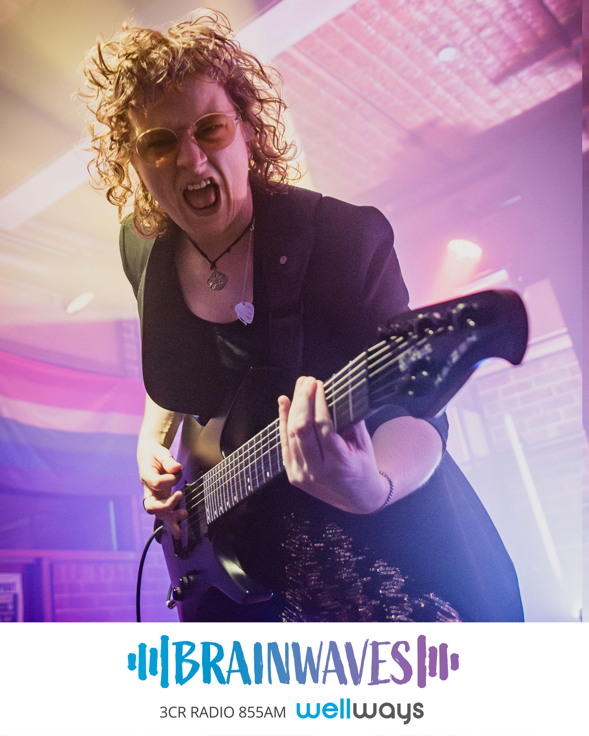 Simone Dow is a red headed caucasian woman on a stage holding and playing a black electric guitar, she wears glasses and a black sequinned jacket and is pulling a screaming face as she leans down towards the camera. There are purple lights behind her.