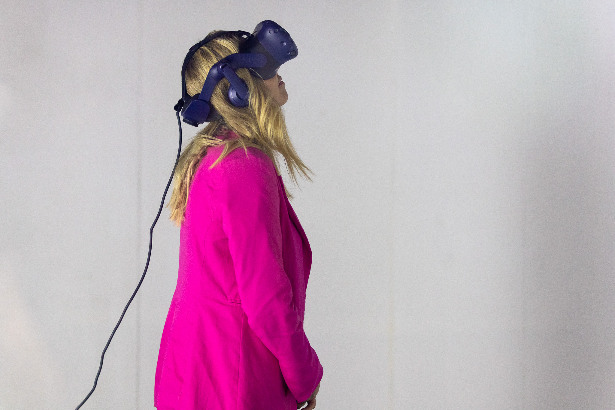 A blonde haired woman in a pink cardigan wears a purple virtual reality headset standing side profile in front of white wall