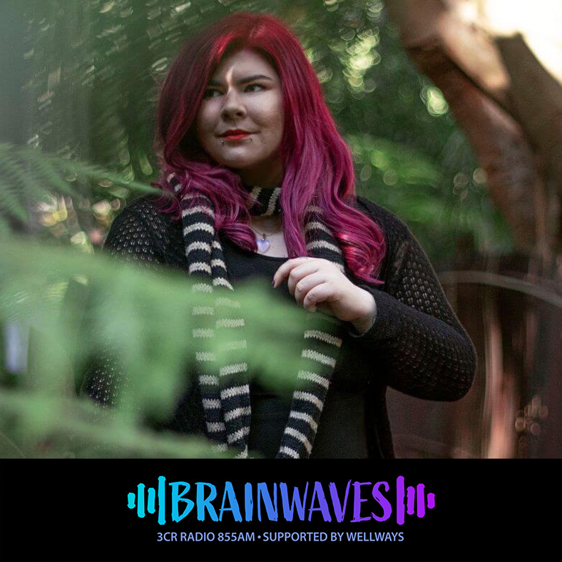 Zoe Simmons is a caucasian woman with long straight pink hair and facial piercings in a nature environment with ferns and green plants surrounding her. She is a Black Summer Bushfire survivor and disabled journalist.