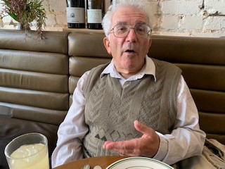 Image of an older man with glasses wearing a cream long sleeved shirt with a colour with a taupe knitted vest over the top. The man is at a cafe sitting in front of him is a beverage and a plate. He looks like he is about to speak. In the background are some wine bottles as decoration.