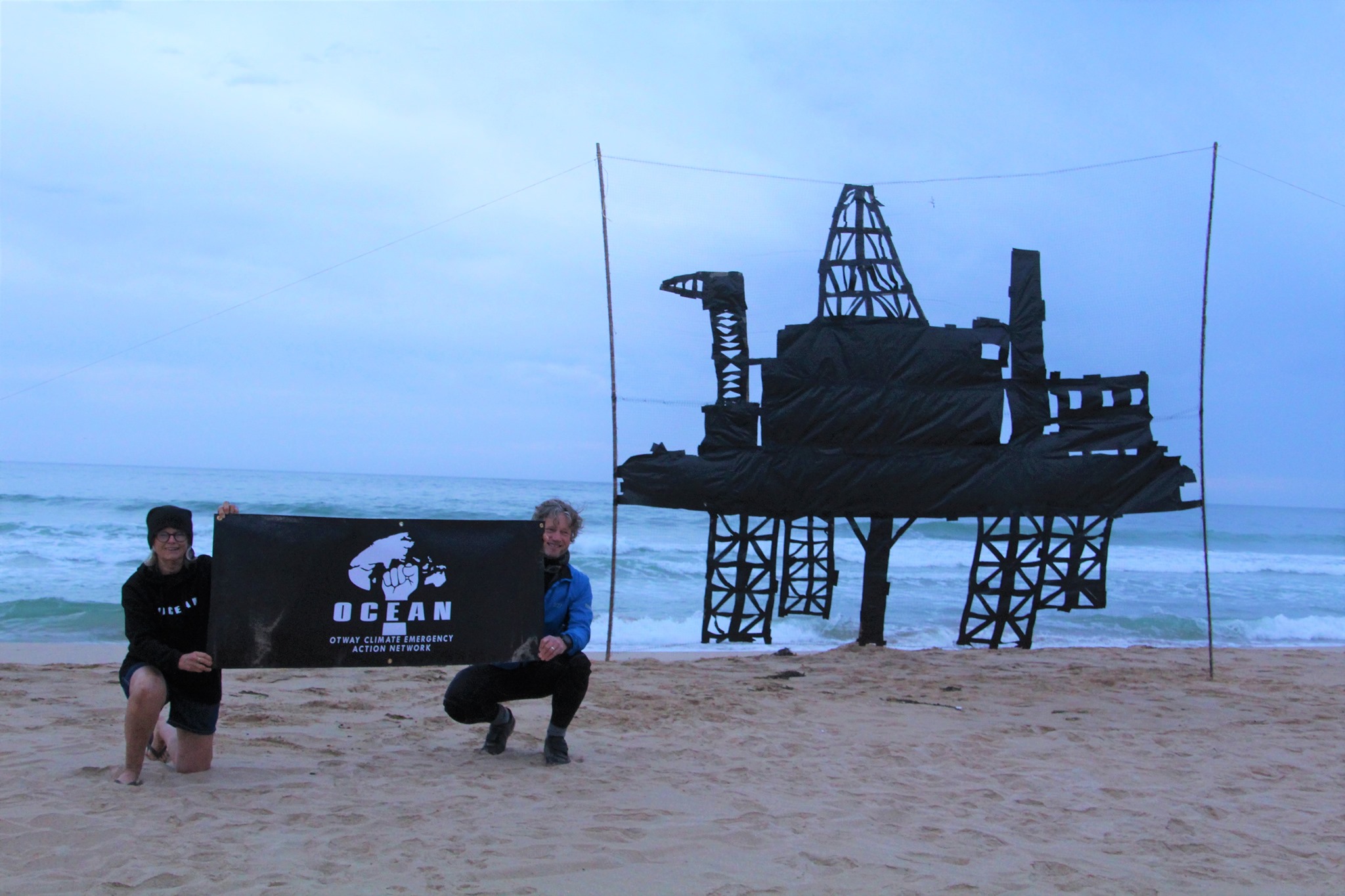 Protest on the beach by OCEAN - black banner with logo and a banner creating the shape of an offshore rig