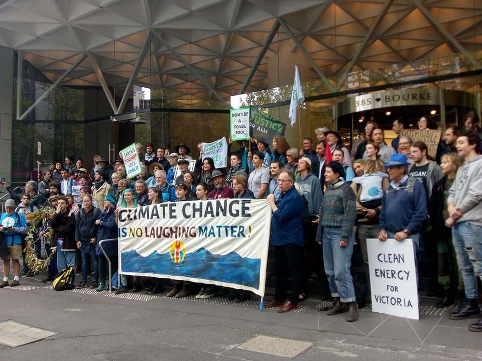 A large group of activists in front of a building holding a "Climate Change is No Laughing Matter" banner