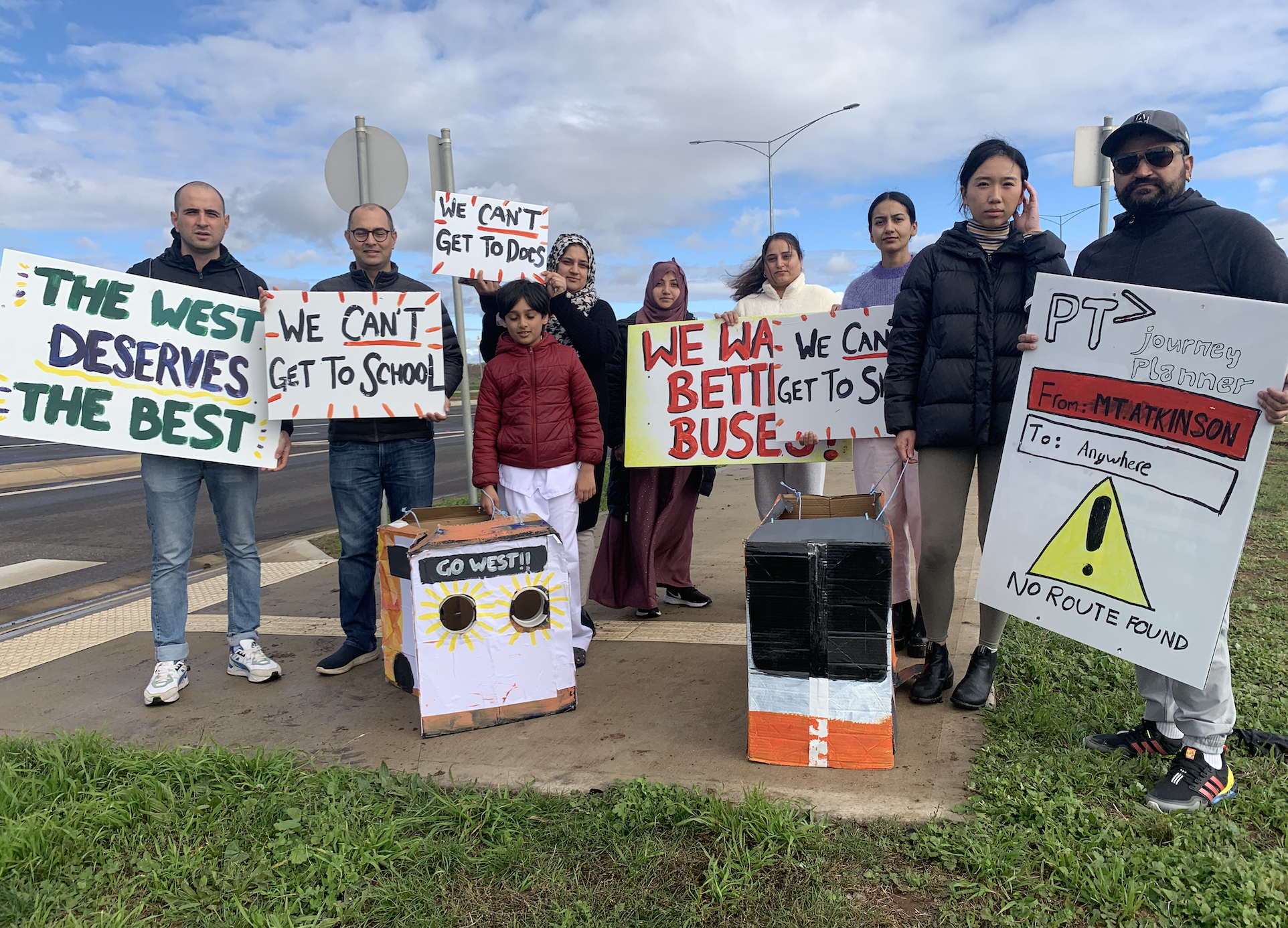 Image of community members from Mt.Atkinson holding signs saying 'Better Buses for the West', 'We can't get to school', 'We can't get to docs', 'We want Better Buses', 'We can't get to shops' and 'PTV journey planner from Mt. Atkinson to Anywhere'