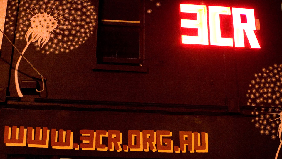 An image of 3CR's building at night time with a mural of a dandelion on the side and large neon sign which reads 3CR, with red text digitally added which reads www.3cr.org.au