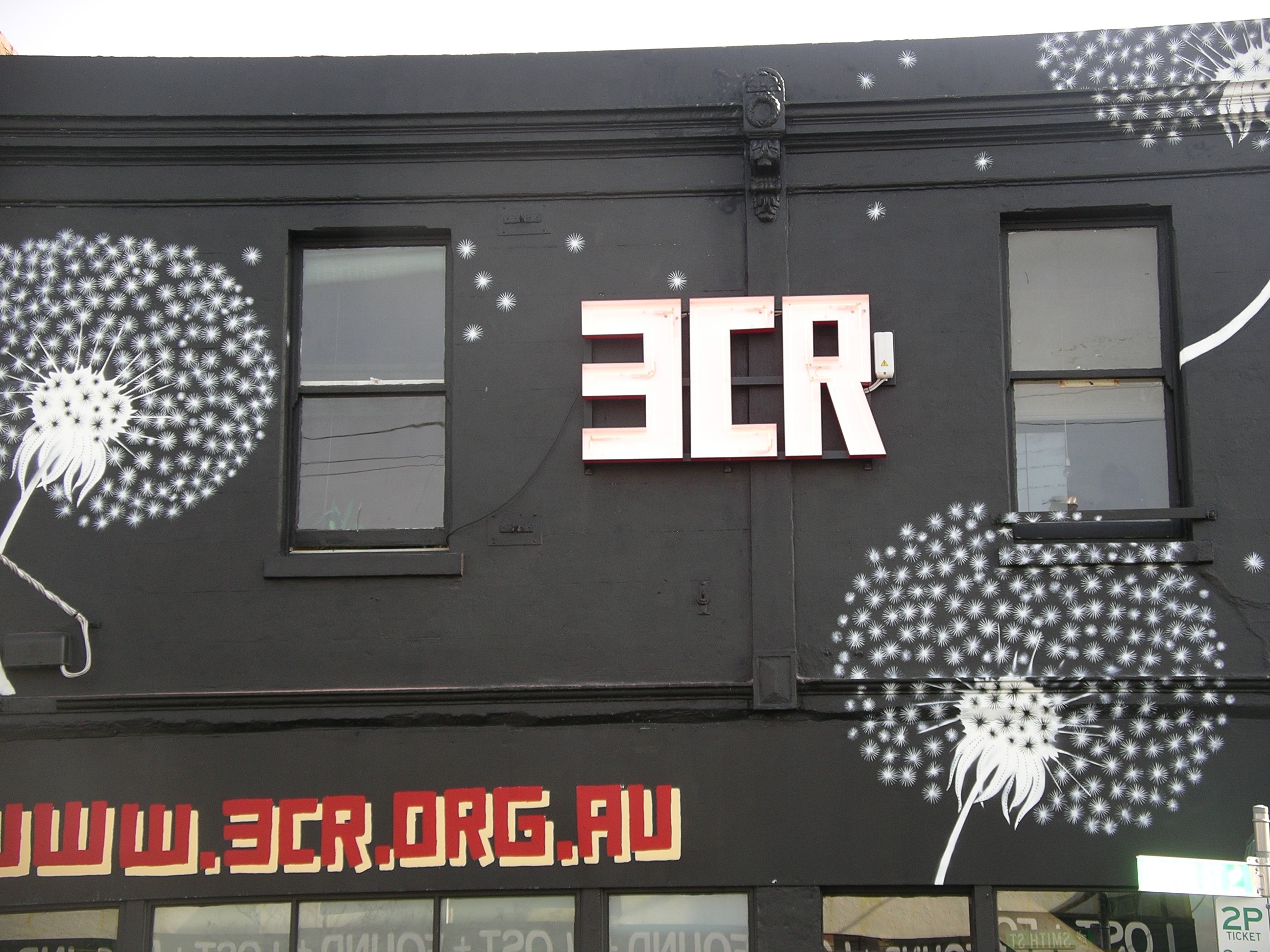 The front of the 3CR building, with large dandelion murals and a neon sign which reads 3CR in block letters.