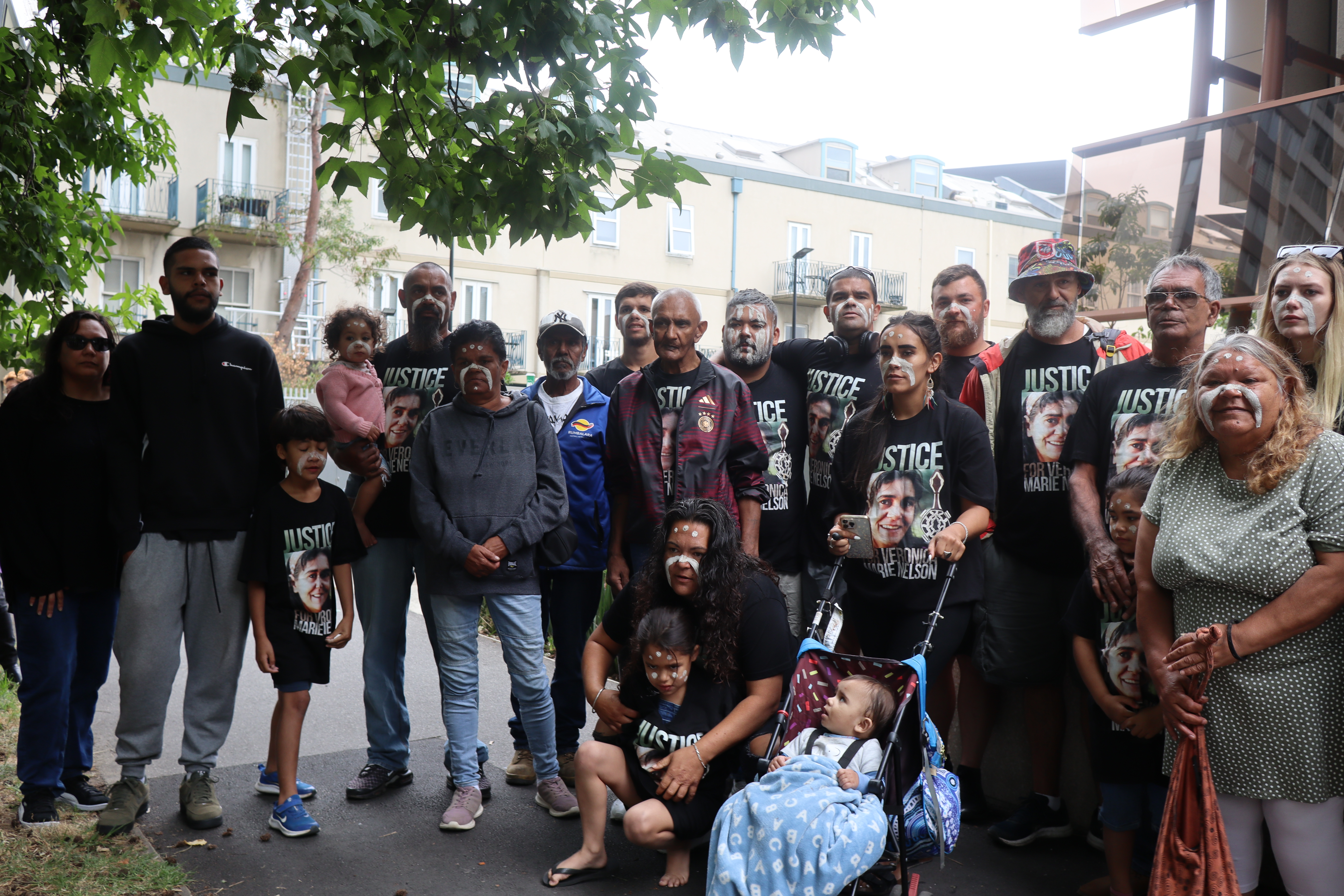 A photograph of 20 of Veronica Nelson's family and friends, including children, looking at the camera and standing together in front of the Coroners Court of Victoria, many of whom are 'painted up' with a curved stripe across nose and cheeks and dots on forehead, and wearing t-shirts that have a photo of Veronica Nelson's smiling face and a line drawing of a turtle that say "JUSTICE FOR VERONICA MARIE NELSON".