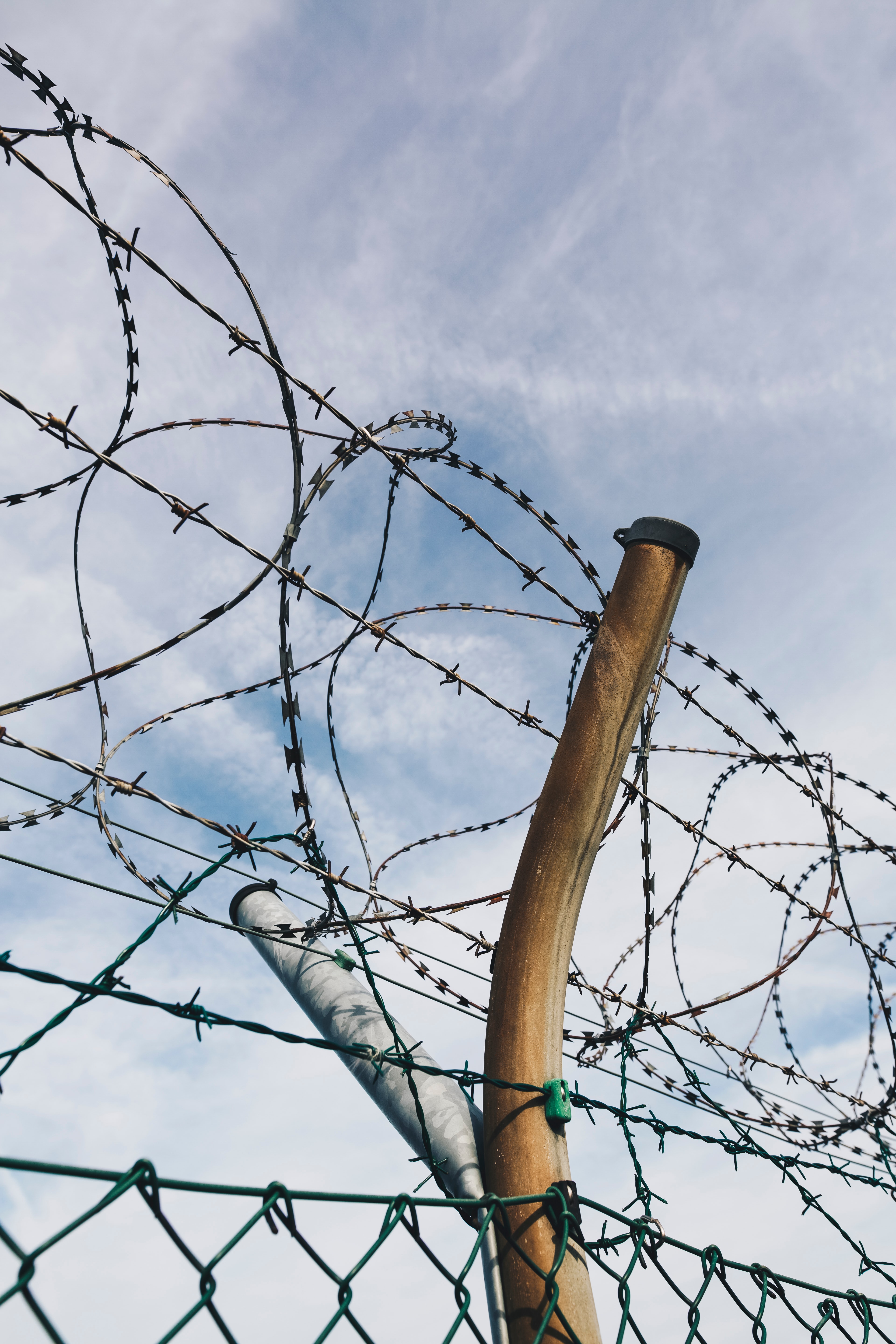 A close-up photograph of a chain-link fence with loops of barbed wire with the sky visible in the background
