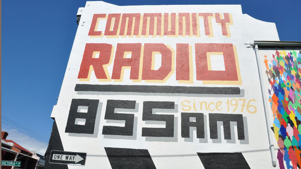 A photograph of the side of the 3CR station building in front of a clear sky. The text painted on the wall reads COMMUNITY RADIO- Since 1976 - 855 AM