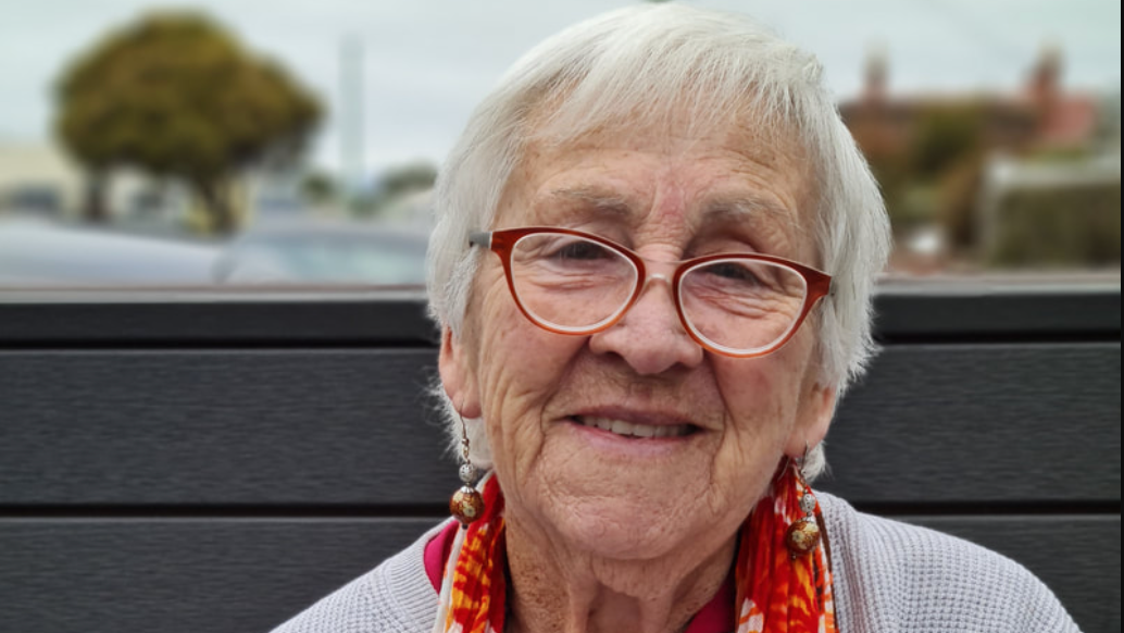 Close-up portrait of the artist. She is an older woman with short silver hair wearing glasses and dangling earings, a colourful orange scarf with a pale blue sweater, and smiling.